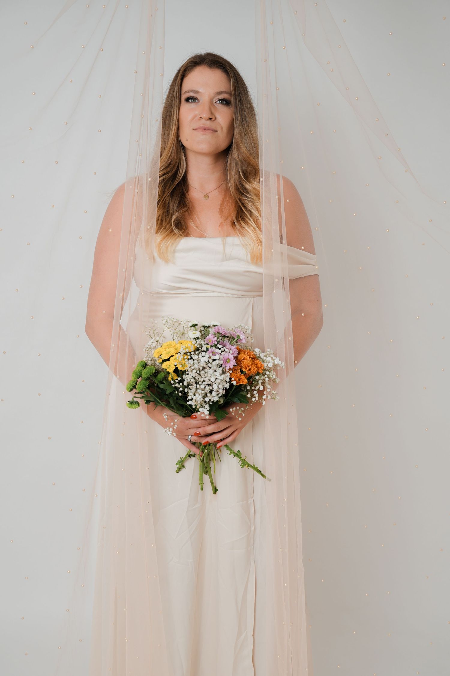 Bride in white gown and veil holding colorful wildflower bouquet, posing against soft draped backdrop.