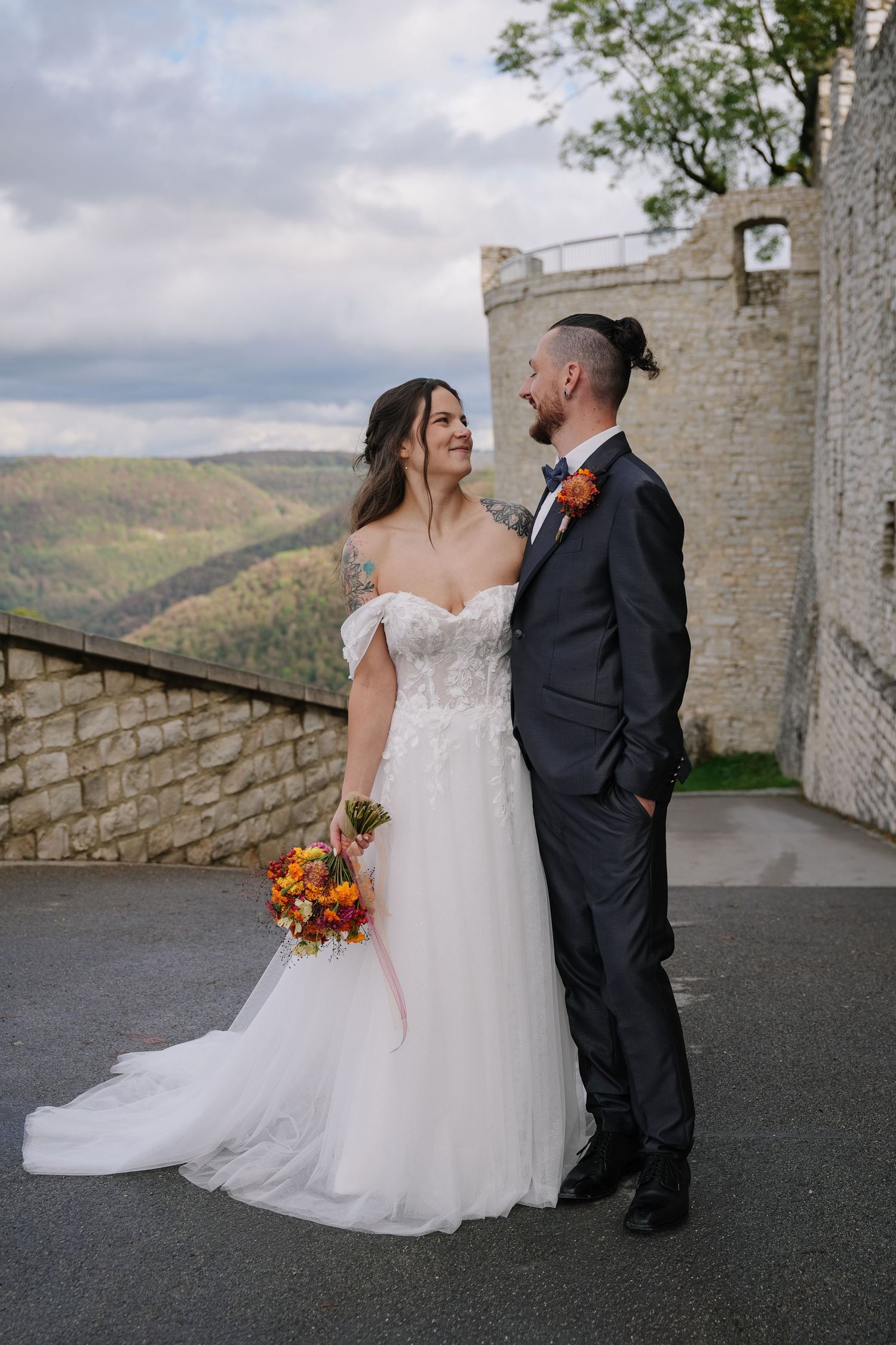 Smiling bride in an off-shoulder lace gown and groom in a navy suit hold an autumn bouquet on a stone terrace overlooking rolling hills.