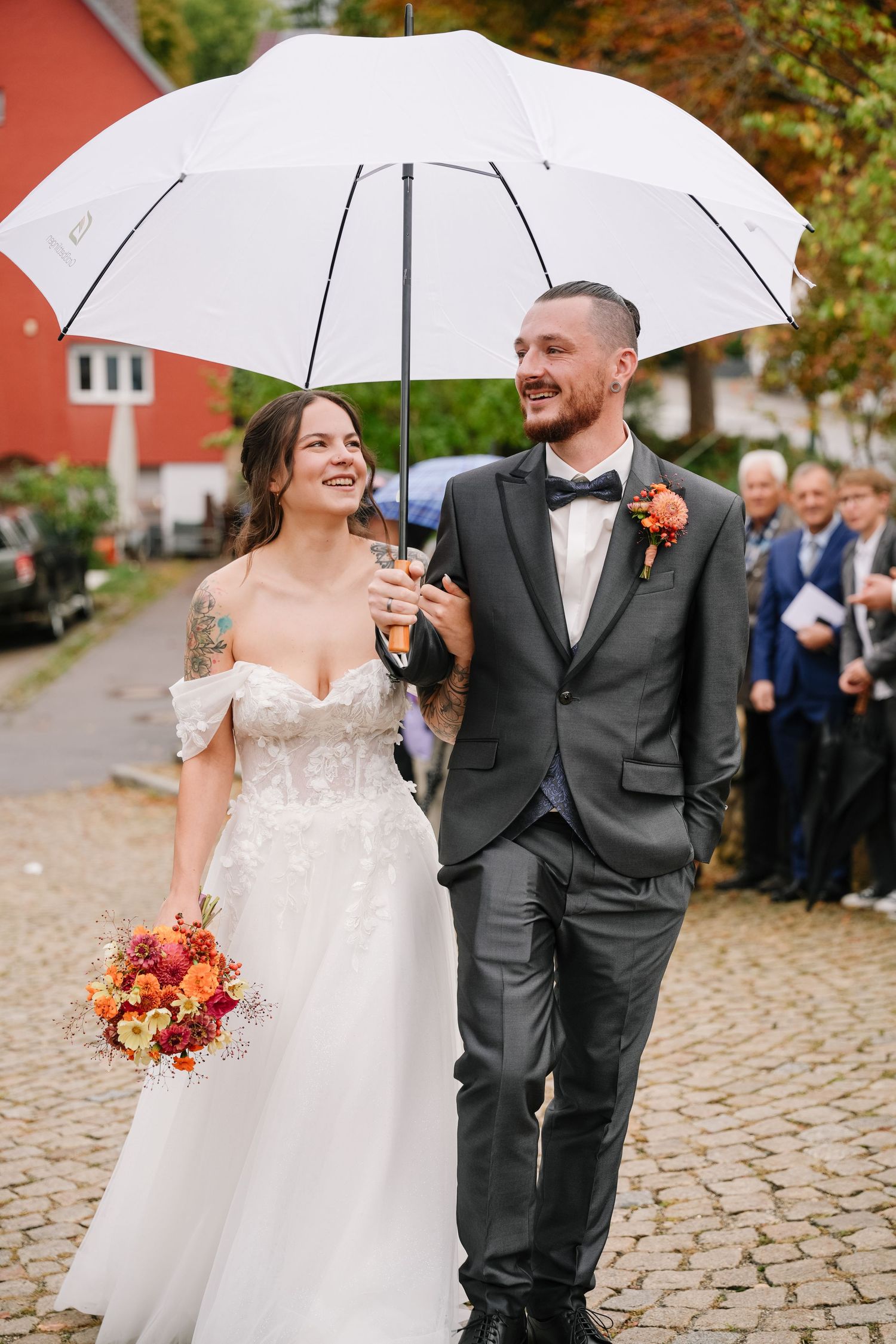 Smiling bride in a lace off-shoulder gown holds an autumn bouquet beside the groom as they walk under a white umbrella.