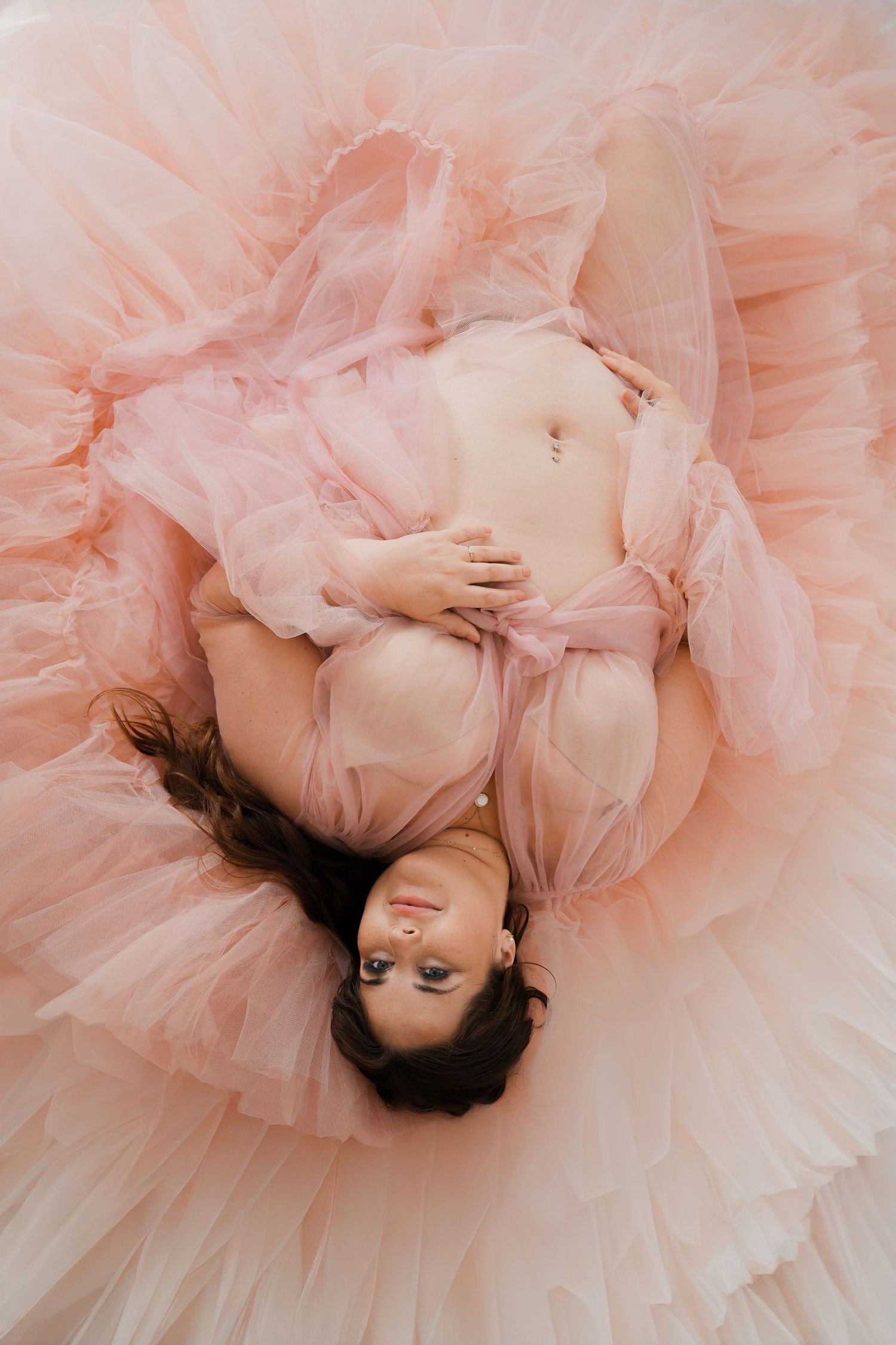 Pregnant woman in pink tulle robe lying down, cradling baby bump, aerial view maternity photography.