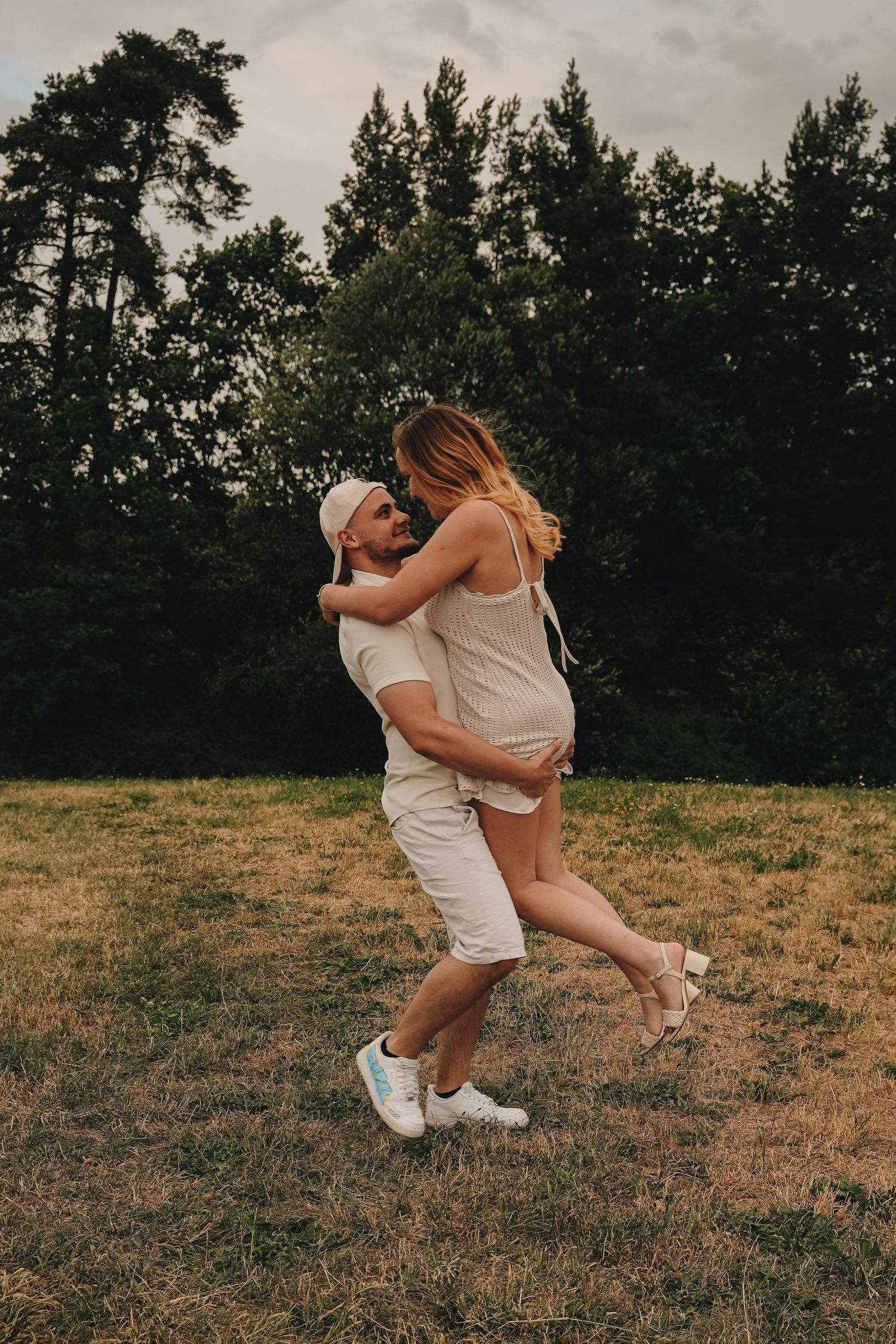 A man joyfully lifts his pregnant partner in a grassy field with lush green trees in the background.