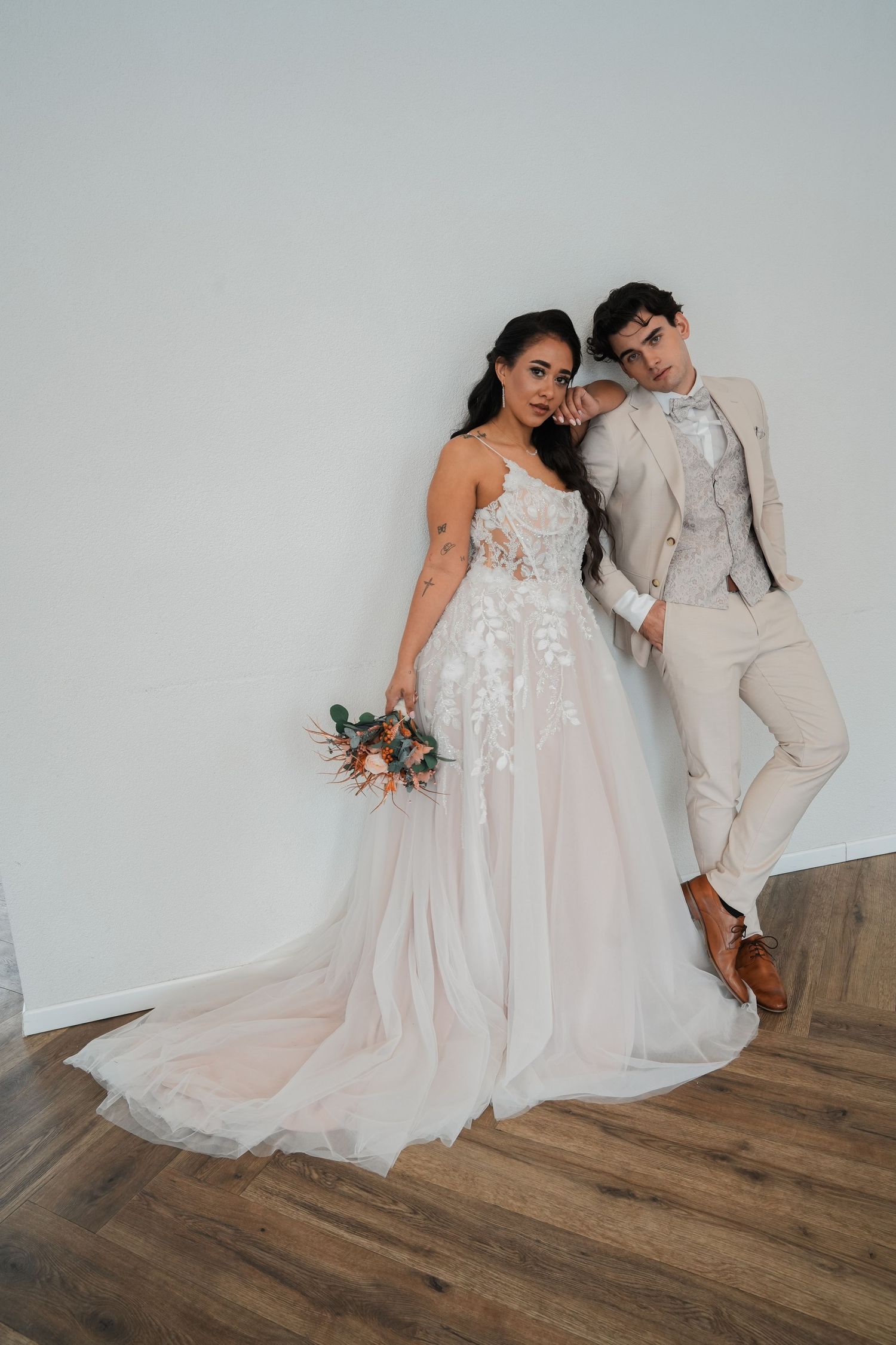 A couple poses against a white wall in elegant wedding attire with a floral bouquet.