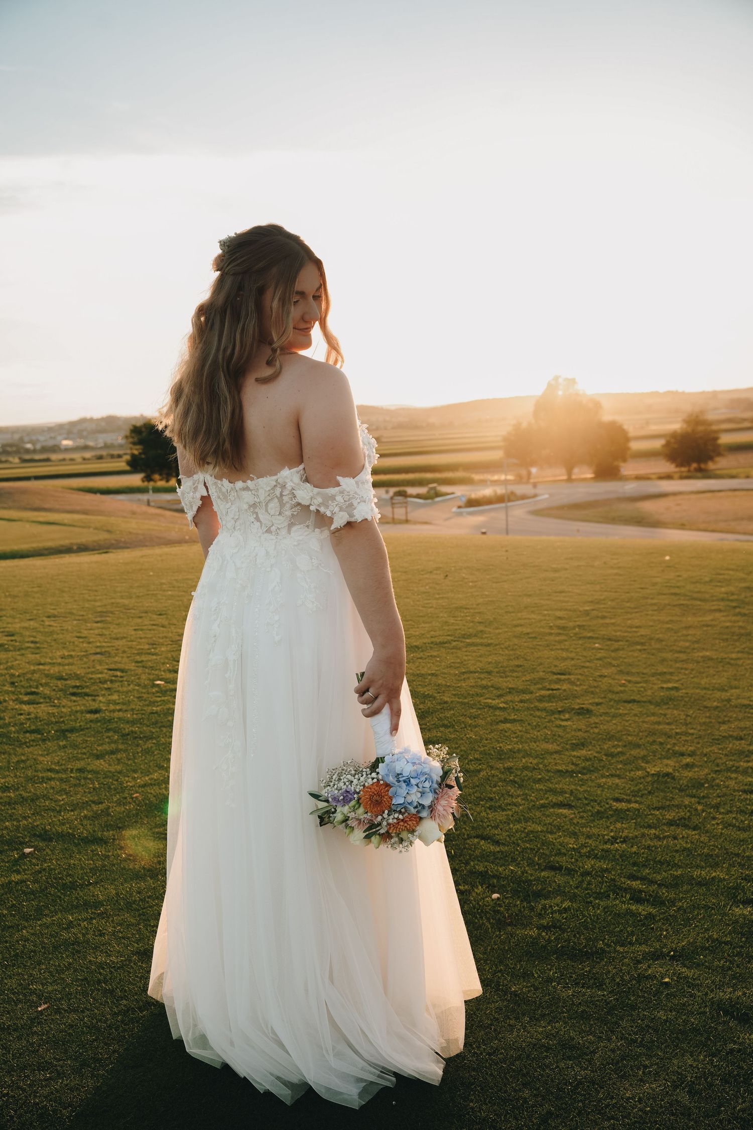Bride in white off-shoulder gown holds floral bouquet at sunset on lush green lawn with scenic countryside backdrop.