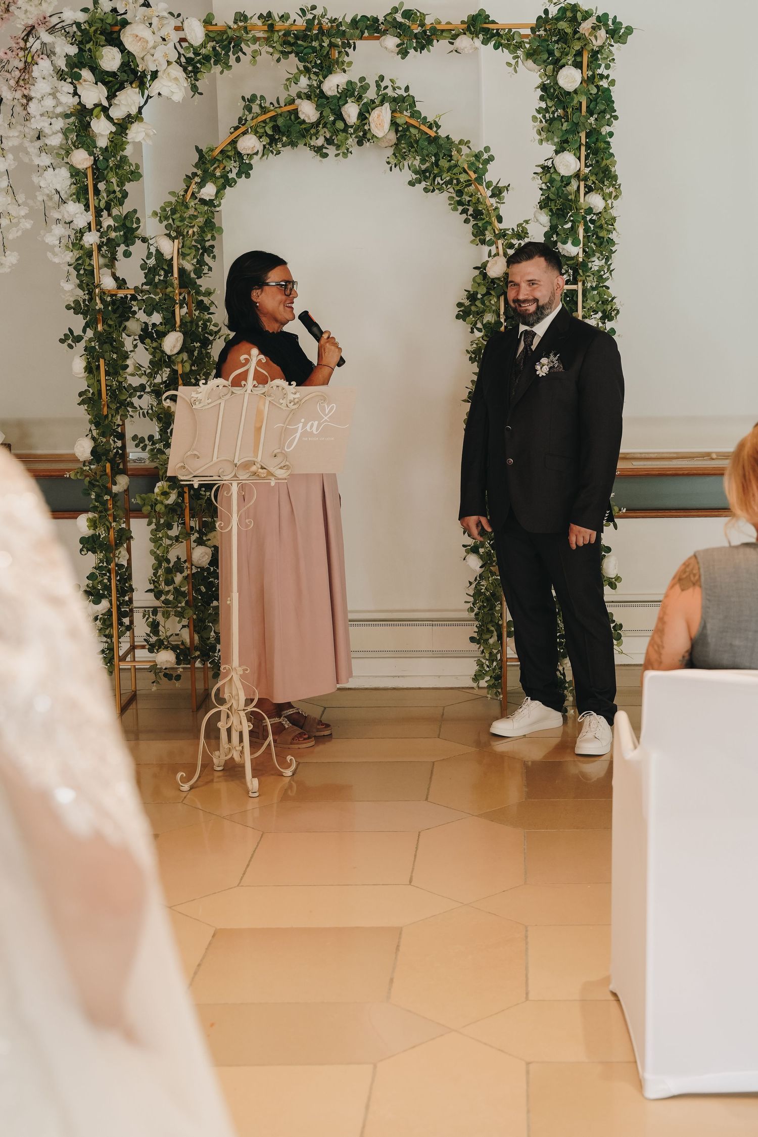 Wedding officiant speaking at a floral arch ceremony with groom in black suit at indoor venue.