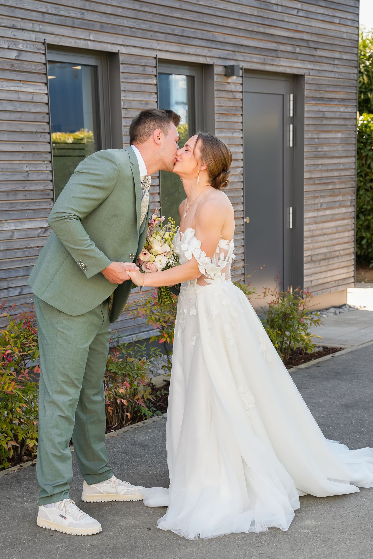 A bride and groom share a kiss outdoors, she in a white gown and he in a sage green suit, holding a bouquet.