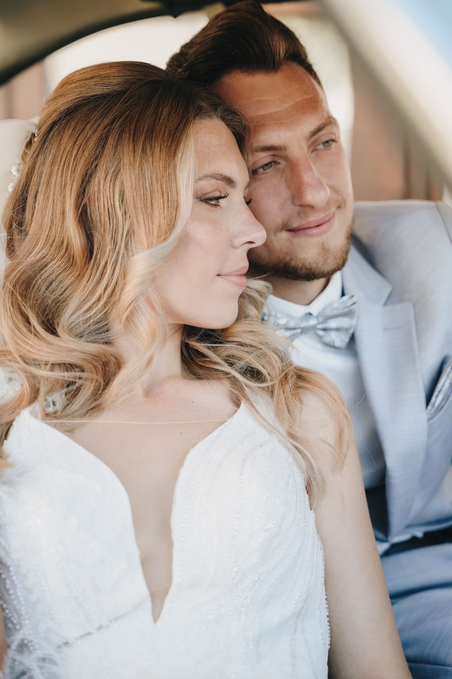 Elegant bride and groom sitting together in a car, wearing white lace dress and gray suit with bow tie.