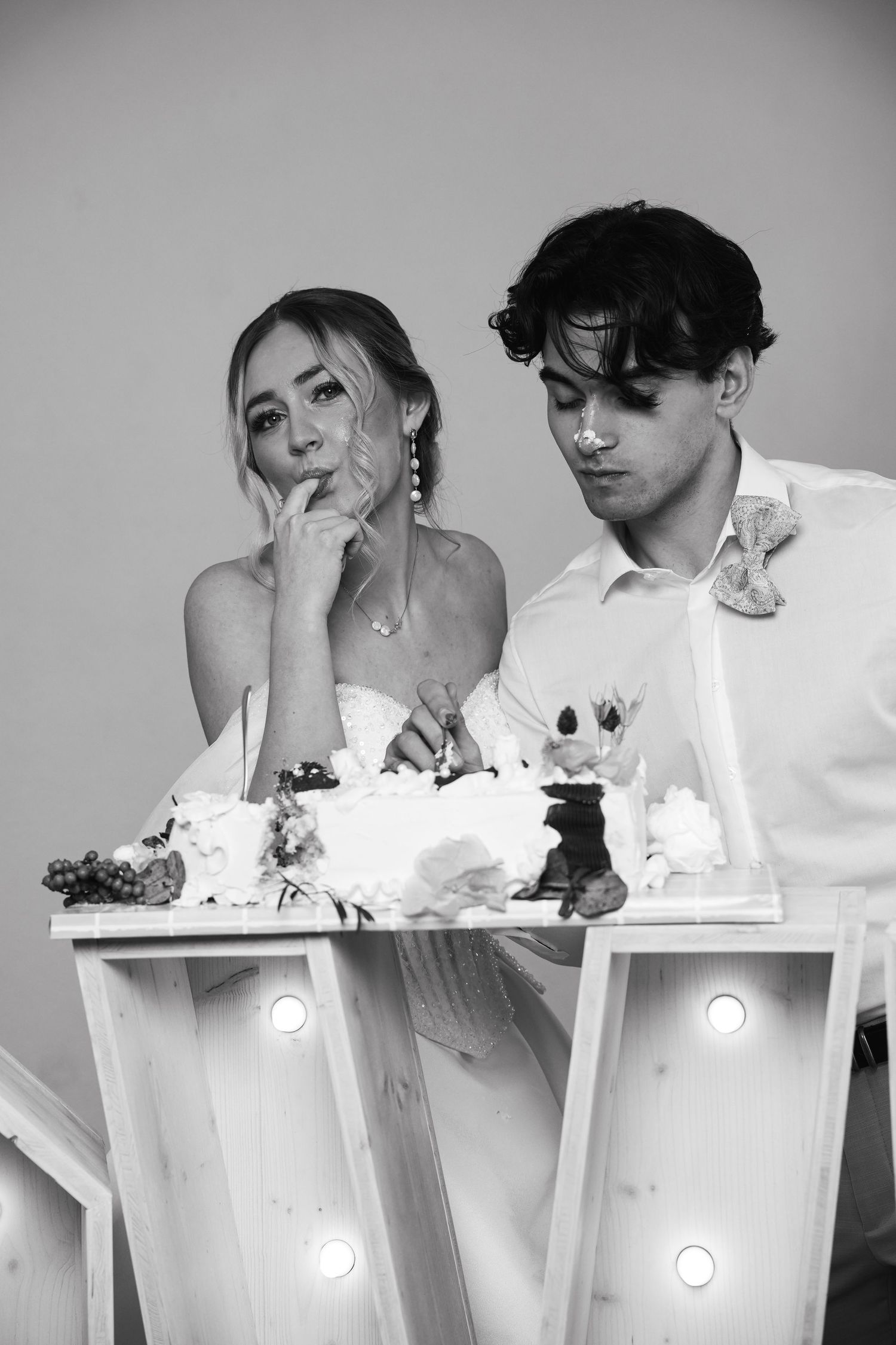 Black and white photo of bride and groom cutting wedding cake on illuminated letter table.