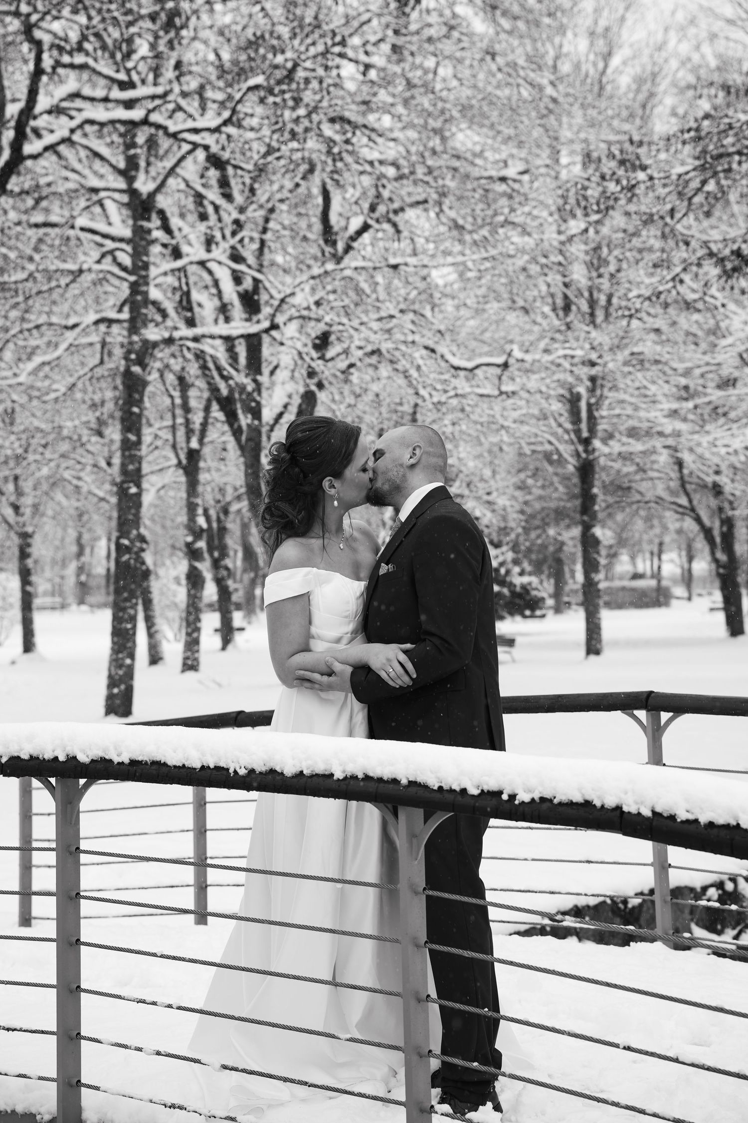 Black and white photo of a bride and groom kissing on a snow-covered bridge surrounded by winter trees.