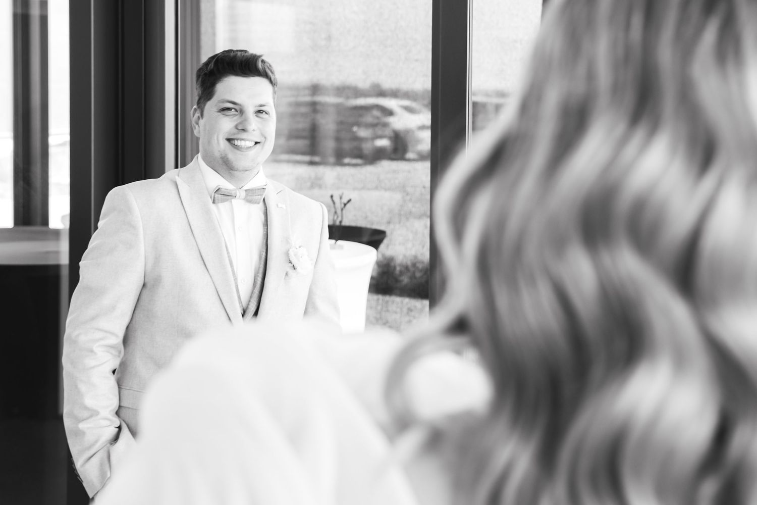 Groom in light gray suit and bow tie smiling at bride during wedding first look moment, black and white photo.
