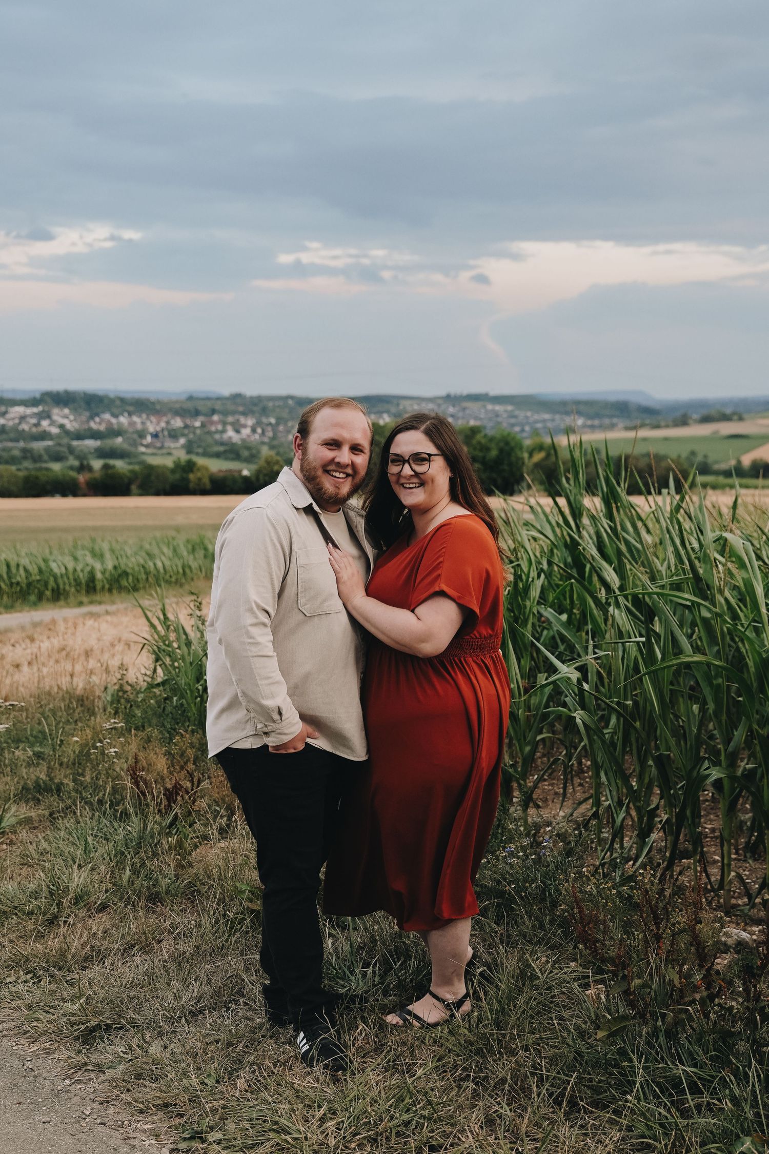 A couple poses together in a red dress and cream shirt by cornfields with a scenic countryside backdrop.