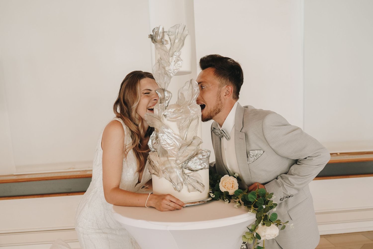 Bride and groom laughing together at a white cake table on their wedding day.