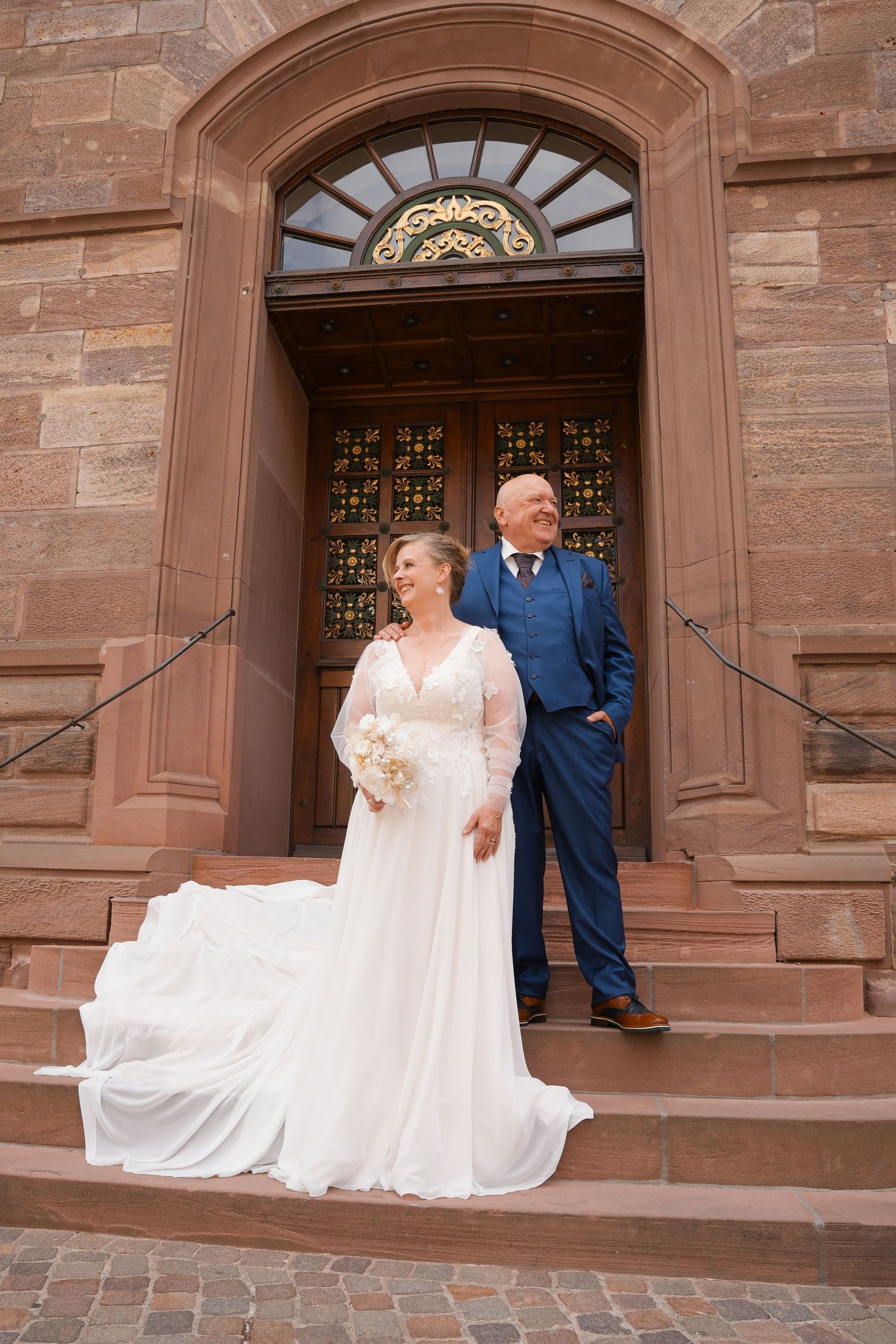Bride and groom stand on church steps, bride in flowing white gown holding bouquet, groom in navy suit.