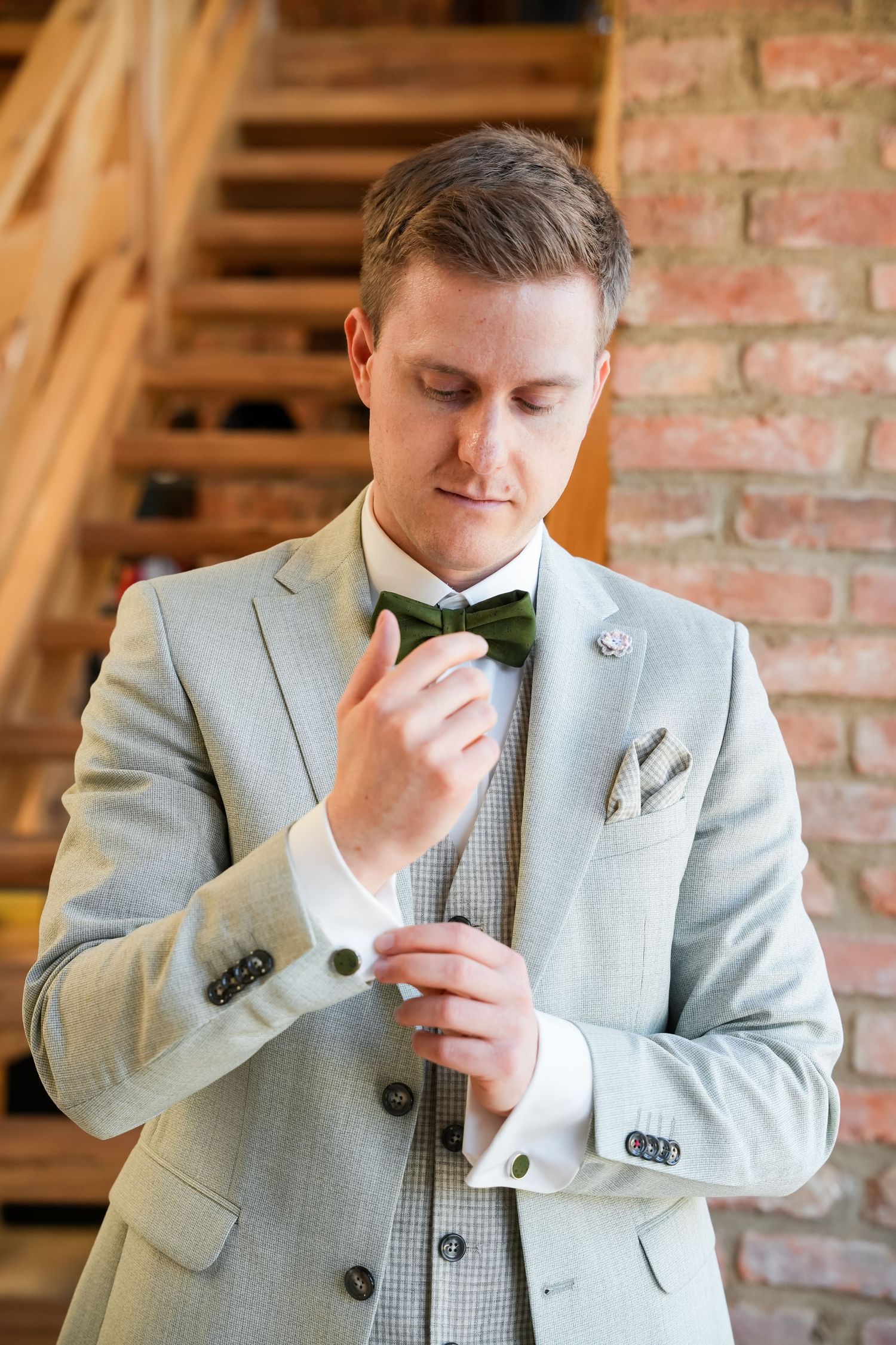 Groom in light gray suit adjusting green bow tie, with brick wall and wooden staircase background.