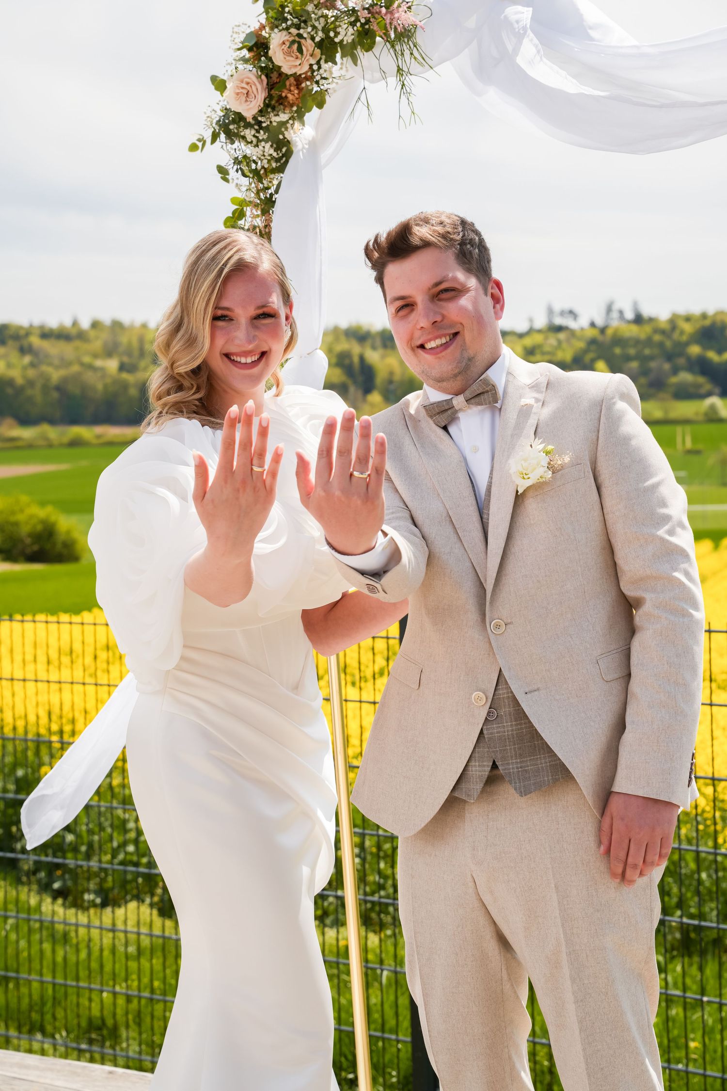 Newlyweds smiling and showing wedding rings at outdoor ceremony with floral arch, bride in white gown, groom in beige suit.