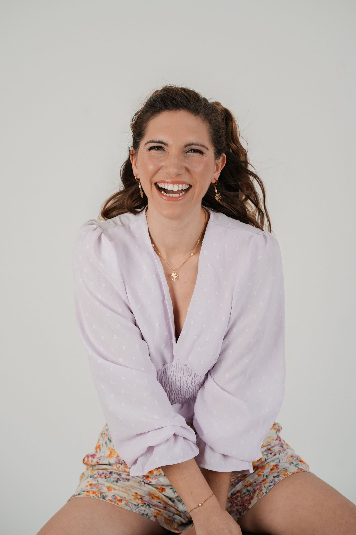 A cheerful woman in a lavender blouse and floral shorts laughs joyfully against a light gray background.