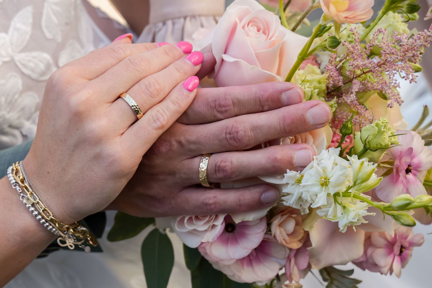 Bride and groom hands with wedding rings resting on a pastel pink rose bouquet.