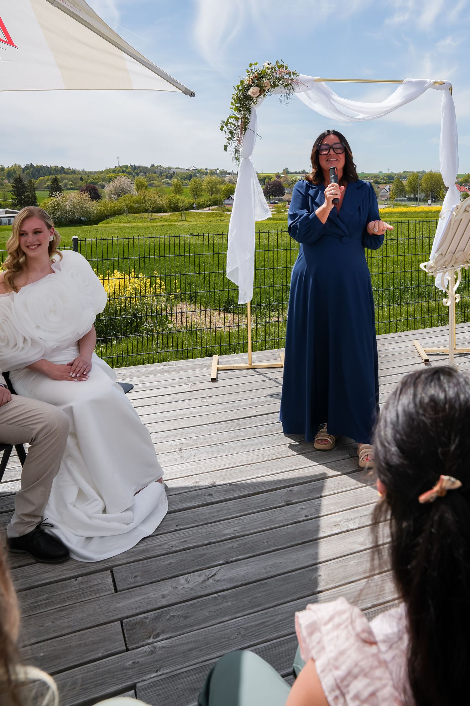 Outdoor wedding ceremony on a deck with bride seated and officiant speaking at floral arch.