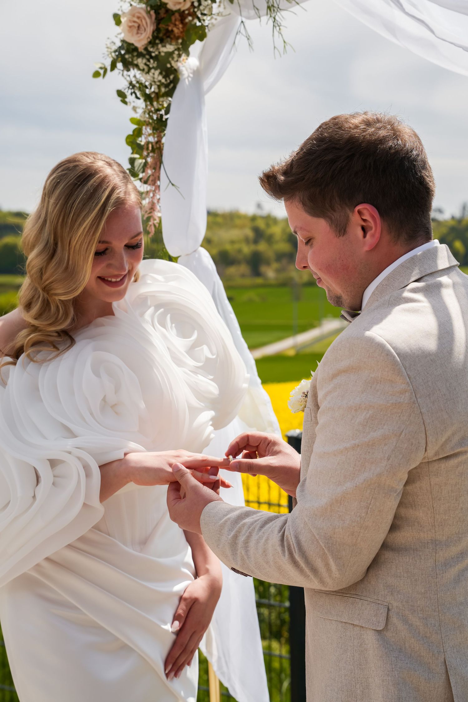 Groom placing wedding ring on bride's finger at outdoor ceremony under floral arch.