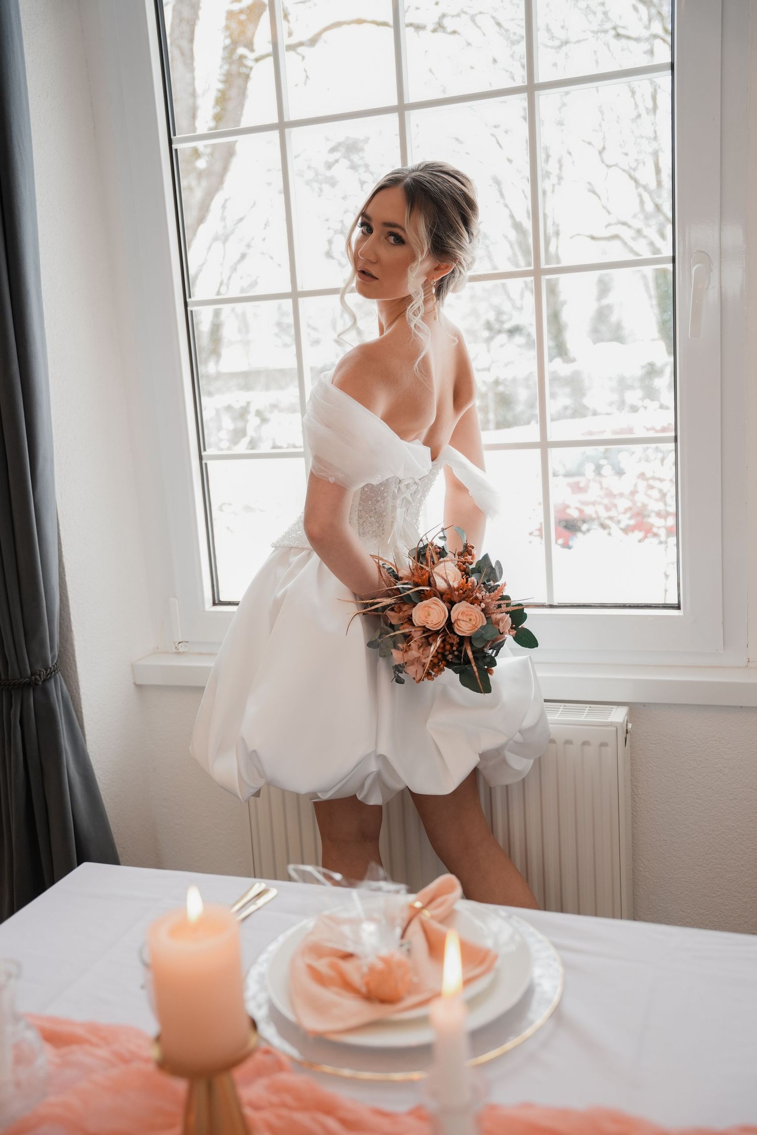 Bride in short white off-shoulder wedding dress holding peach bouquet stands by window with snowy view and candlelit table.
