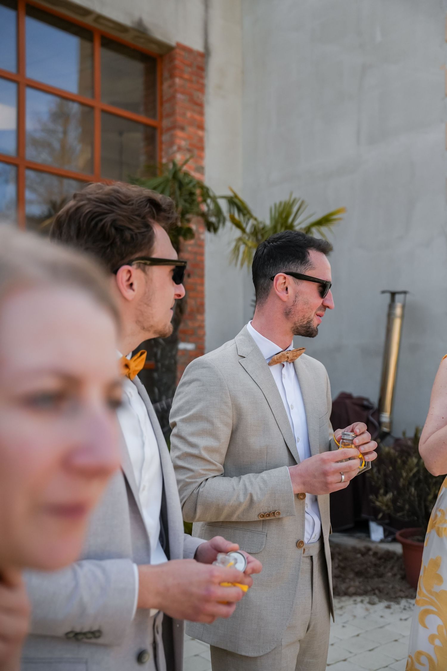 Men in tan suits and bow ties with sunglasses socializing outdoors at a wedding reception.