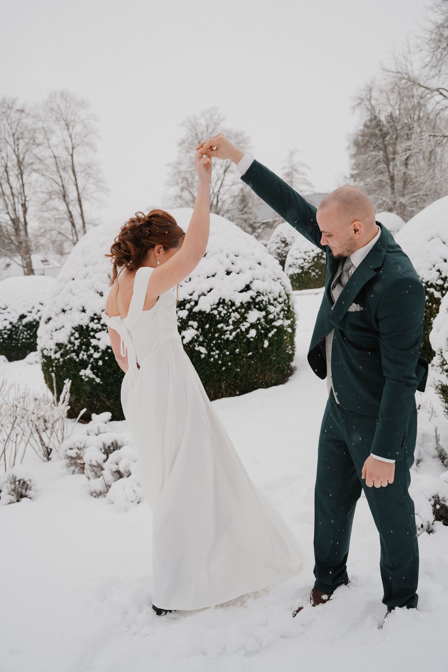 Bride and groom dancing in snowy winter garden, she wears white gown, he wears dark green suit.