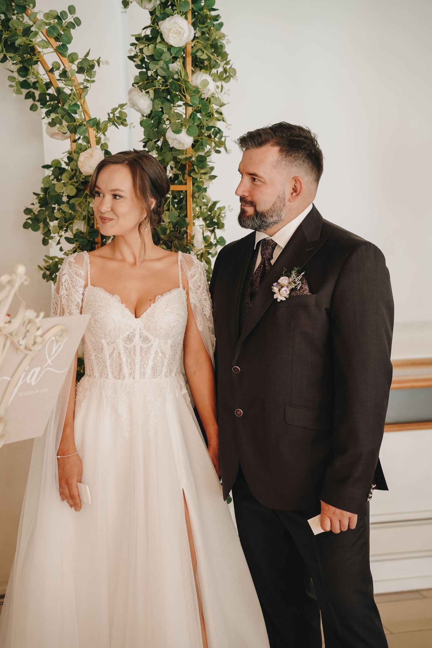 Bride and groom holding hands at wedding altar with white floral arch and greenery backdrop.
