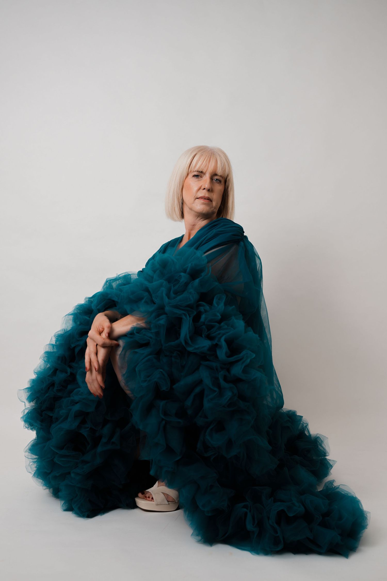 Middle-aged woman with blonde bob sits on floor wearing dramatic teal ruffled tulle gown against white background.