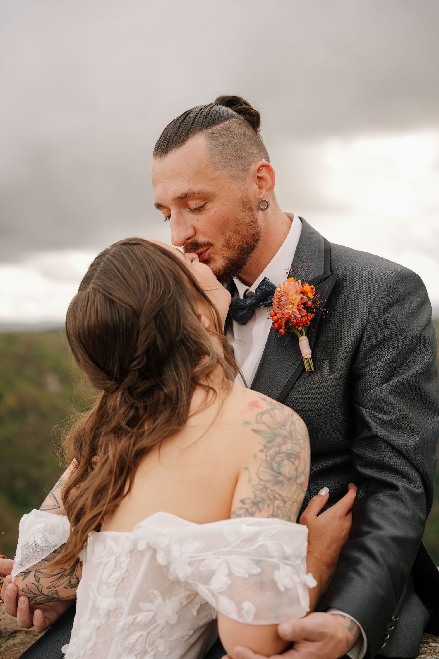 Groom in a gray suit with orange boutonniere kisses his bride in an off-shoulder lace gown against a cloudy, scenic overlook.