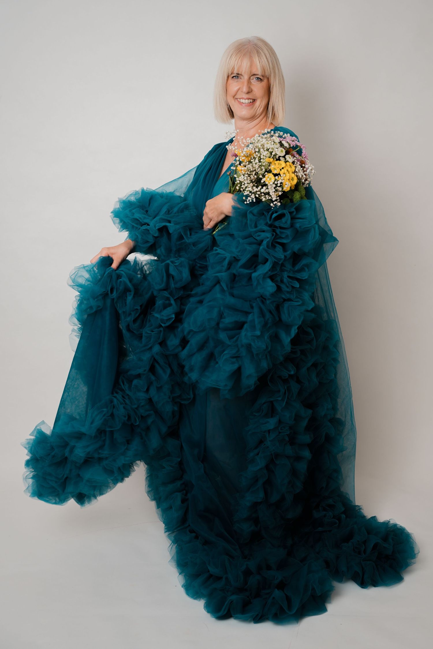 Elegant older woman in teal ruffled gown holding wildflowers, posing gracefully against white background.