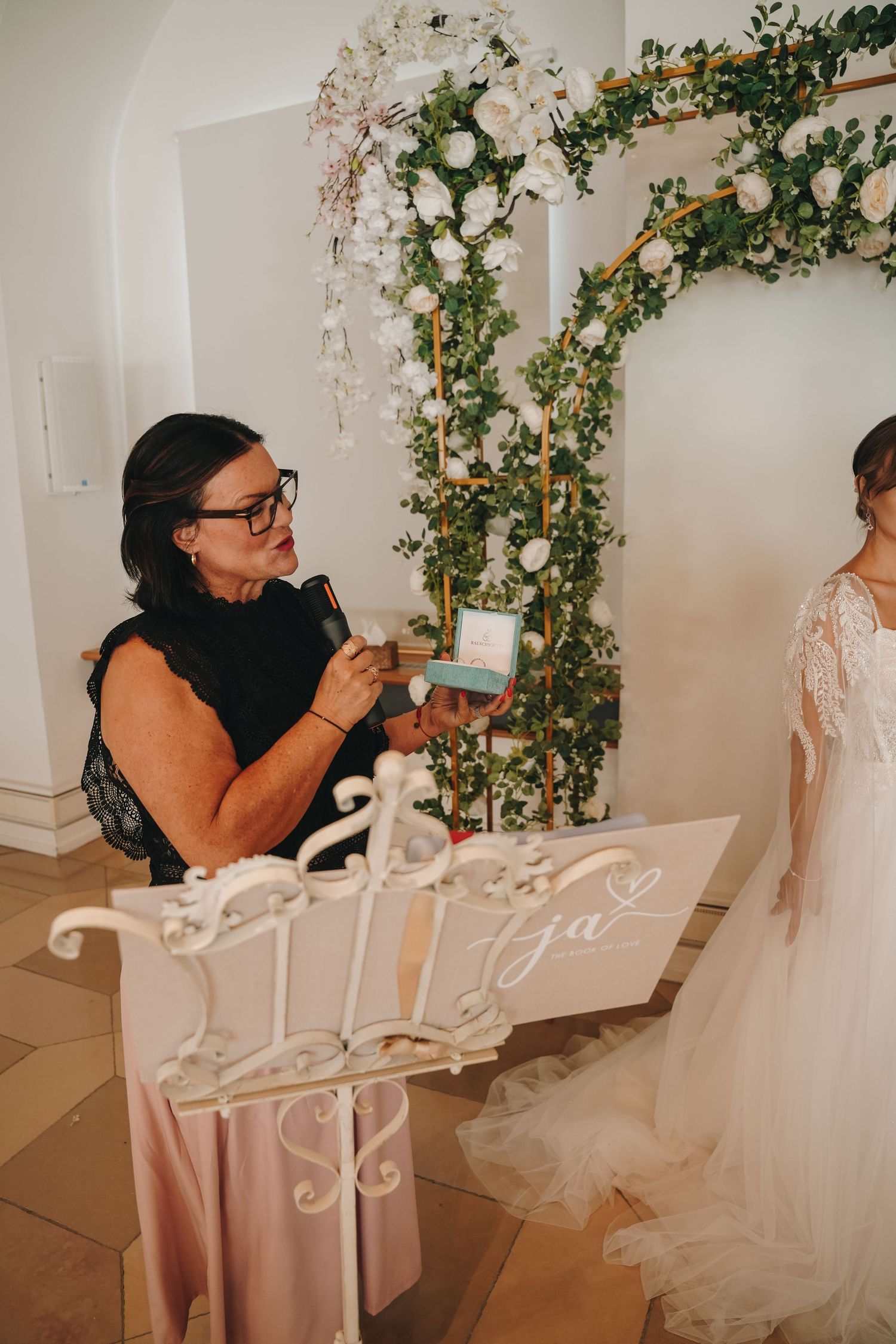 Wedding officiant holding ring box at floral arch ceremony, bride in lace gown stands nearby at elegant indoor venue.