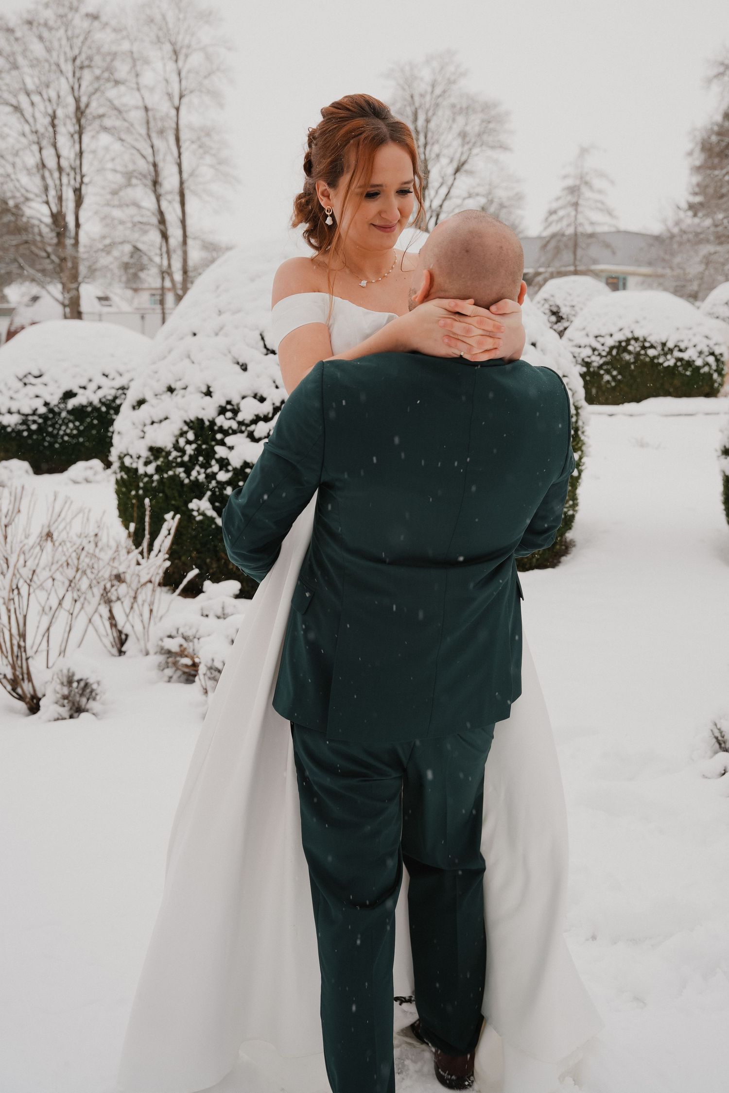 Bride in white off-shoulder gown embraces groom in green suit during snowy winter wedding outdoors.