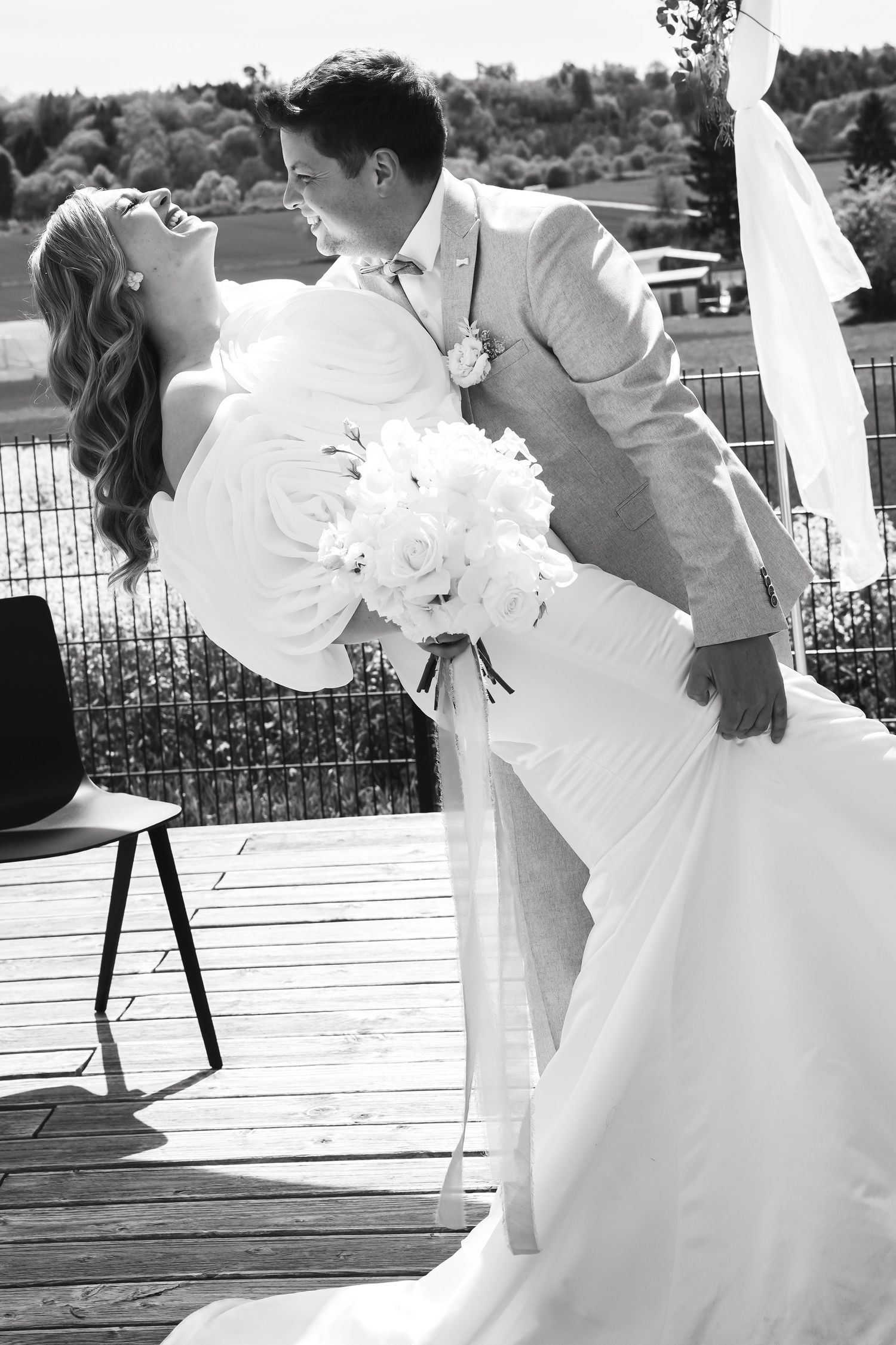 Black and white photo of groom dipping bride in wedding dress holding bouquet on outdoor deck.