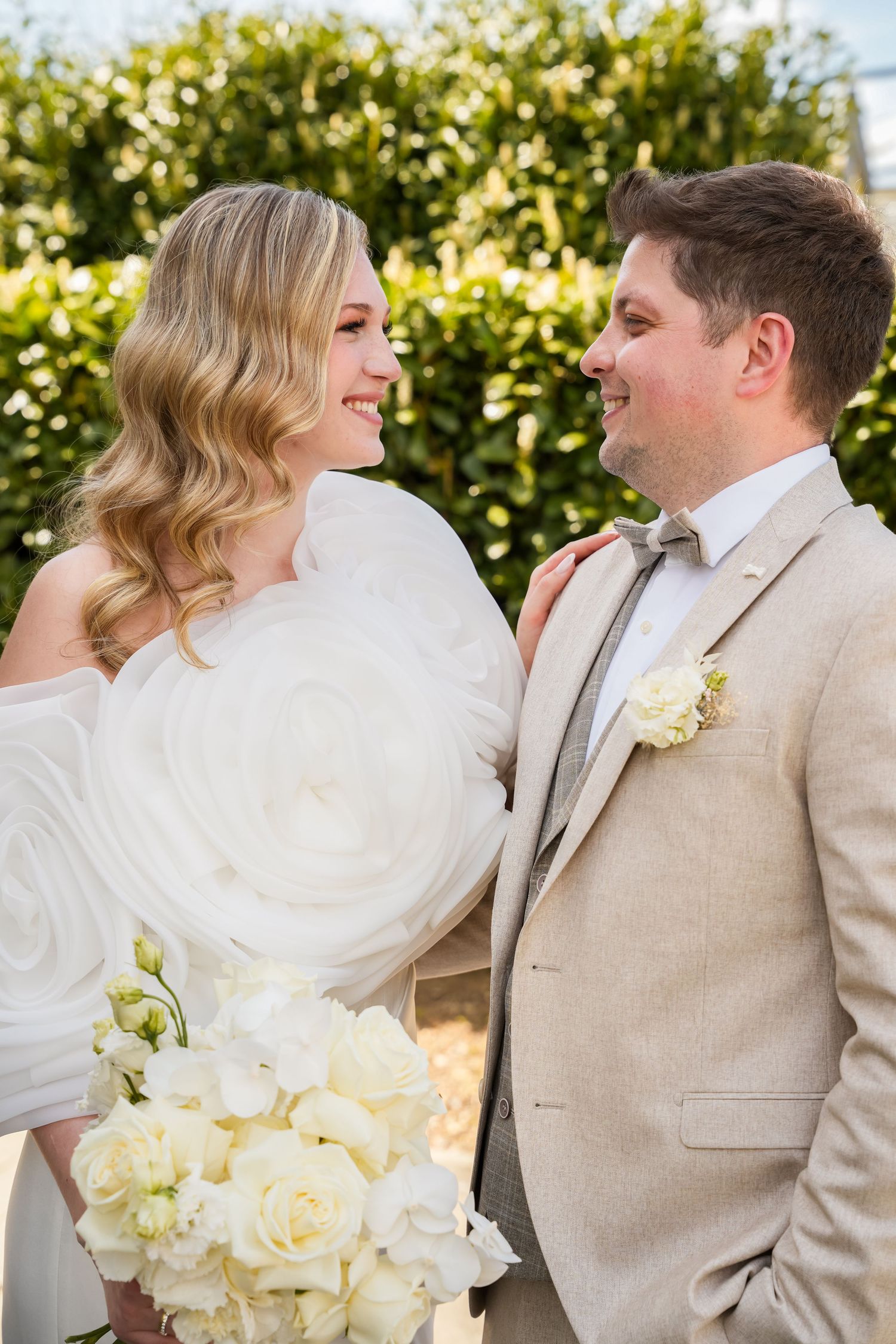 Smiling bride in white off-shoulder gown holding white roses bouquet stands with groom in beige suit outdoors.