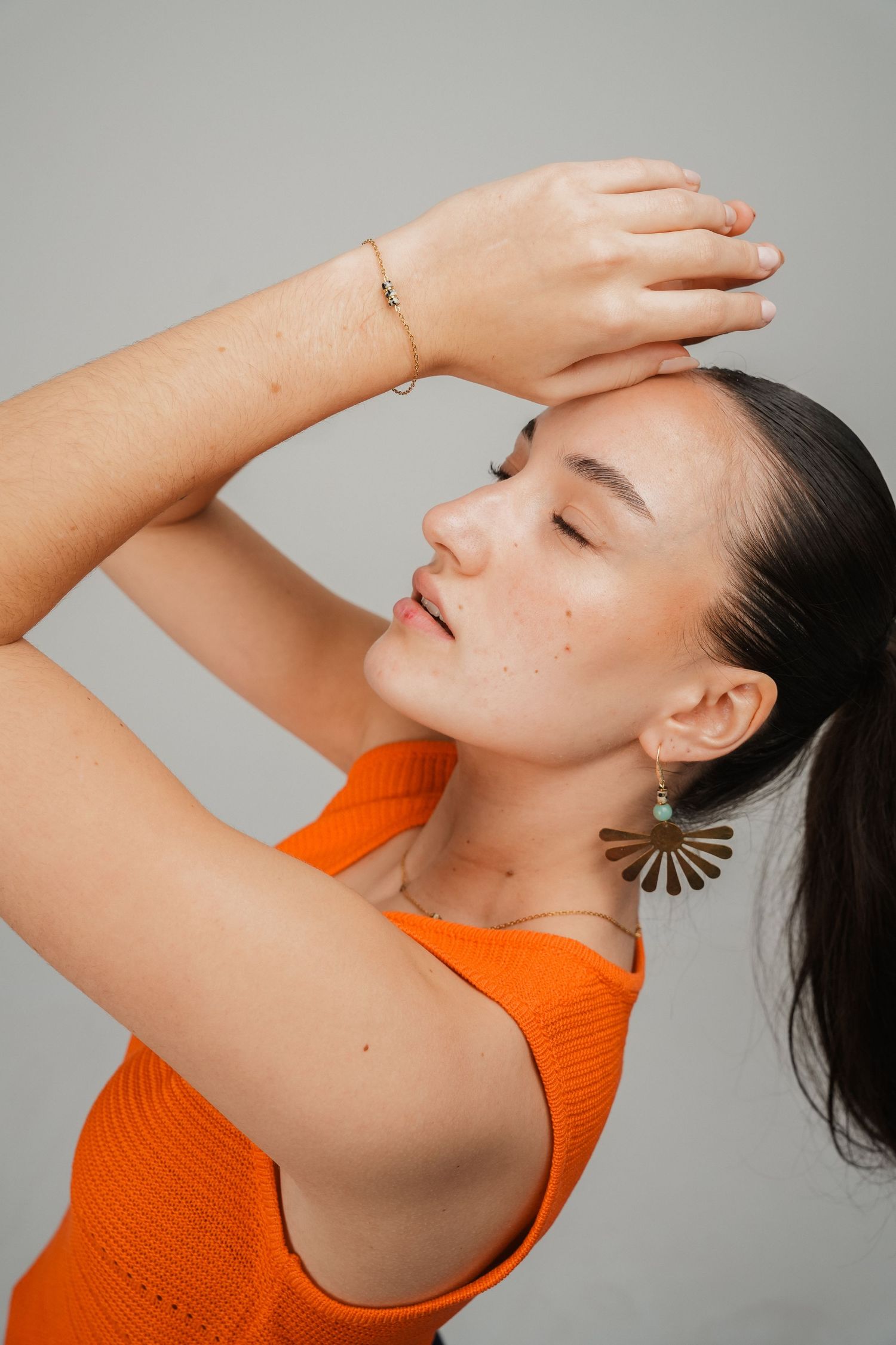 Woman in orange top poses with hands raised, wearing gold fan earring and bracelet against gray background.