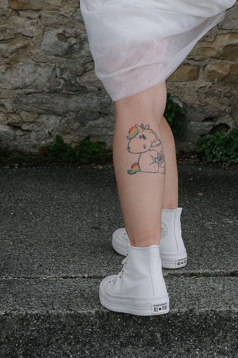 Person in white high-top Converse sneakers and a white skirt stands on stone steps showing a rainbow unicorn tattoo on the calf.