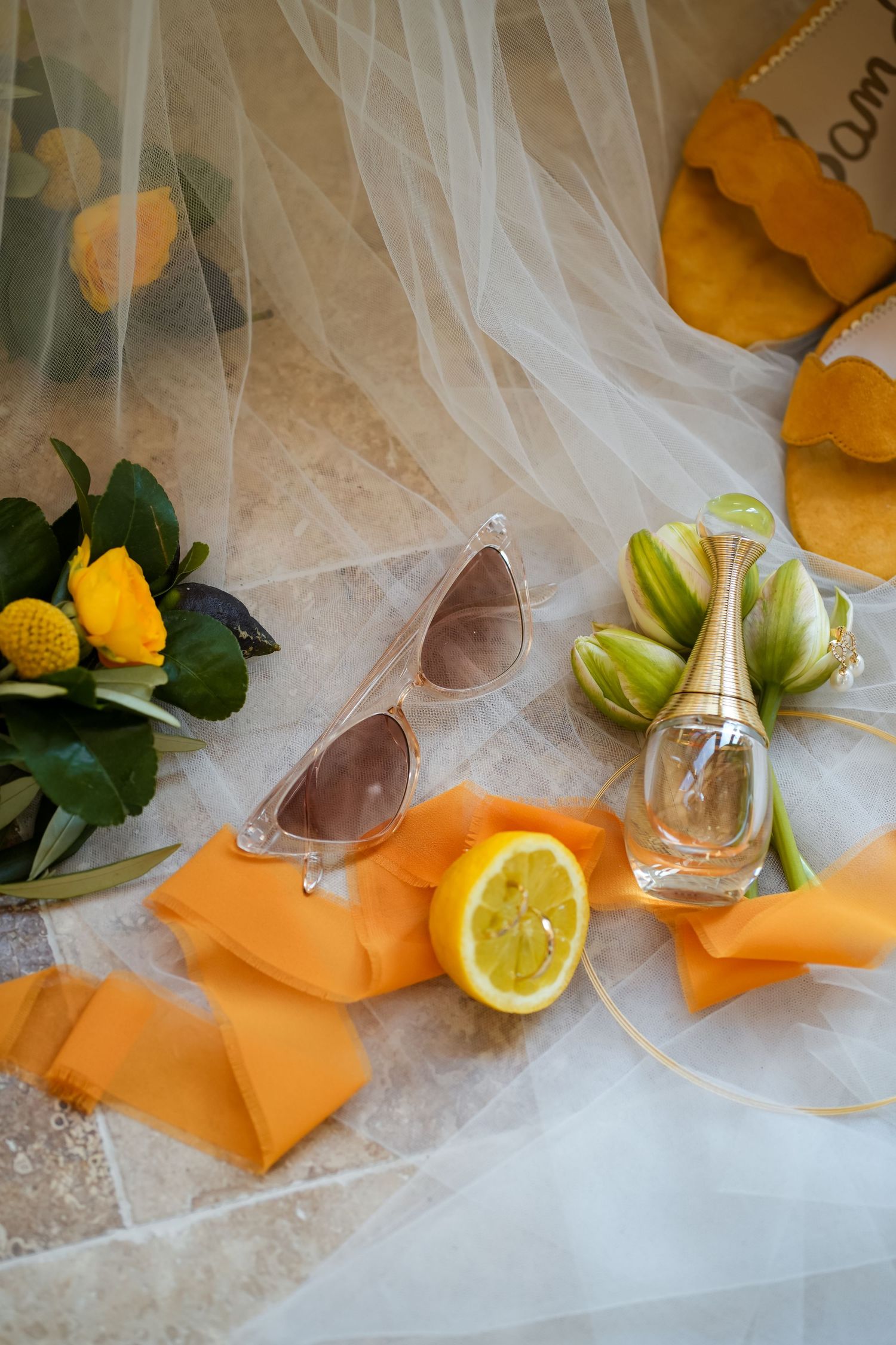 Bridal flat lay with sunglasses, Dior perfume, lemon, orange ribbons, yellow roses, and gold sandals on white tulle.