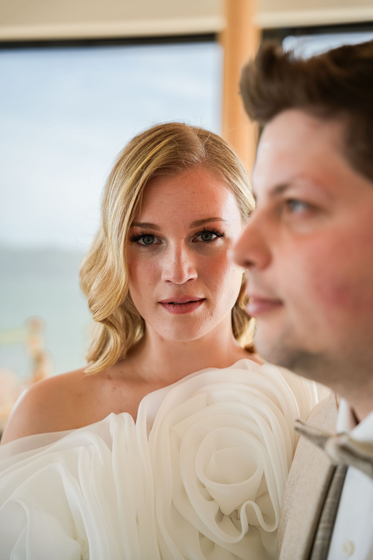 Bride in white ruffled gown gazes at groom during wedding ceremony with ocean view backdrop.