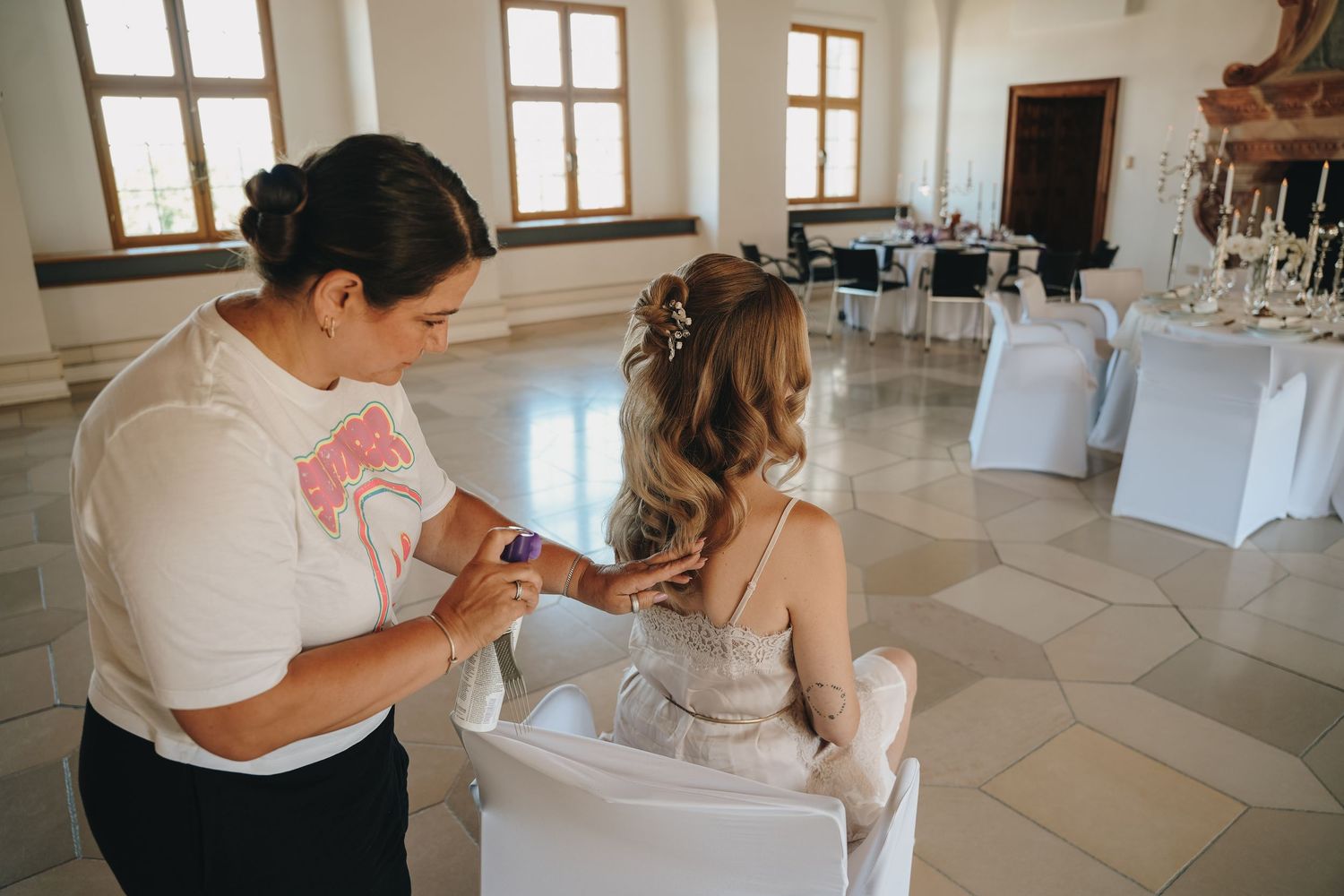 Hairstylist adjusting bride's elegant updo hairstyle in a decorated wedding venue hall.