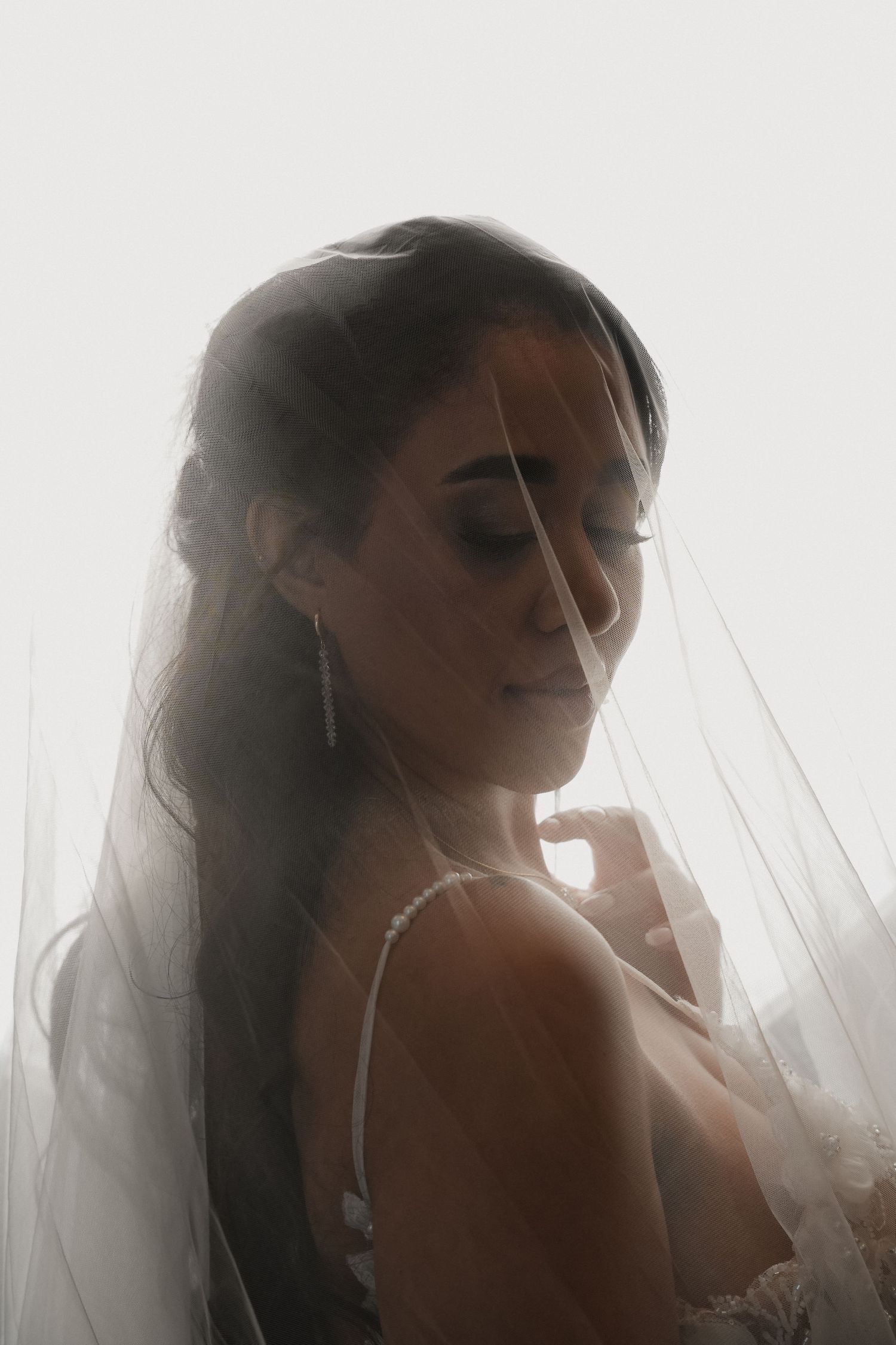 Bride in white lace gown draped in a sheer veil, looking down gracefully in soft backlit light.