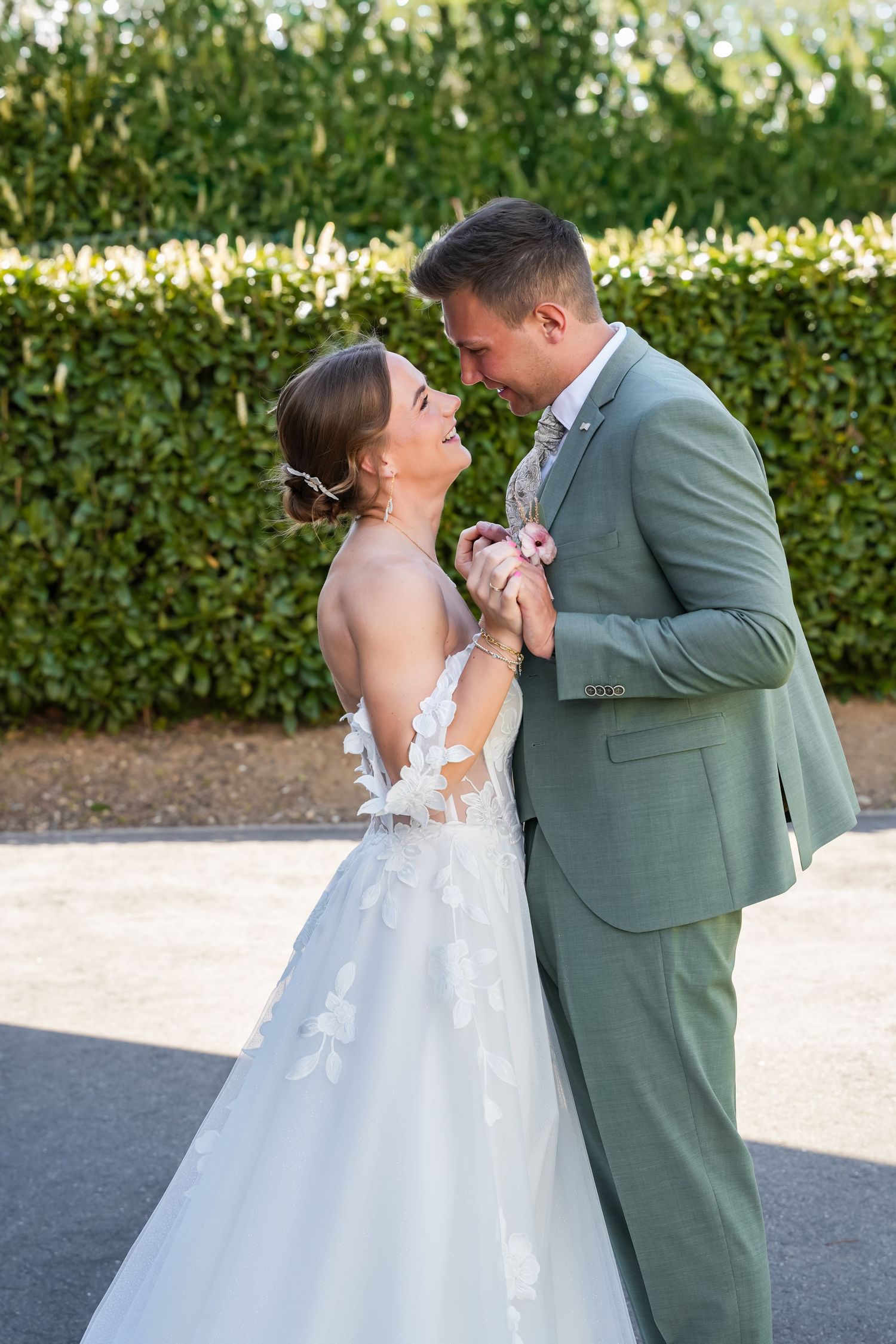 A bride in a floral lace gown and groom in a sage green suit share a romantic moment outdoors near a lush hedge.