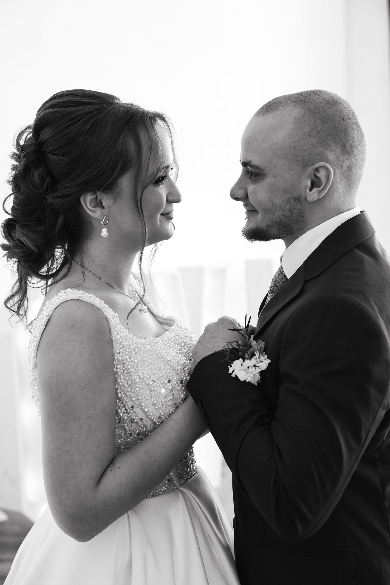 Black and white wedding photo of smiling bride and groom gazing at each other, bride in pearl dress, groom in dark suit.