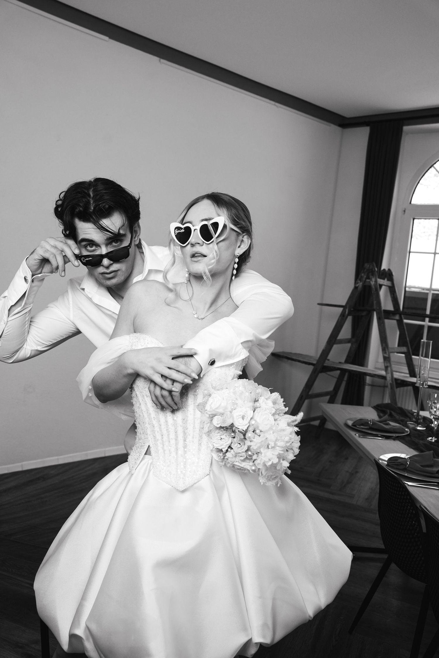 Black and white photo of a playful bride and groom wearing sunglasses, bride holding bouquet in elegant white dress.
