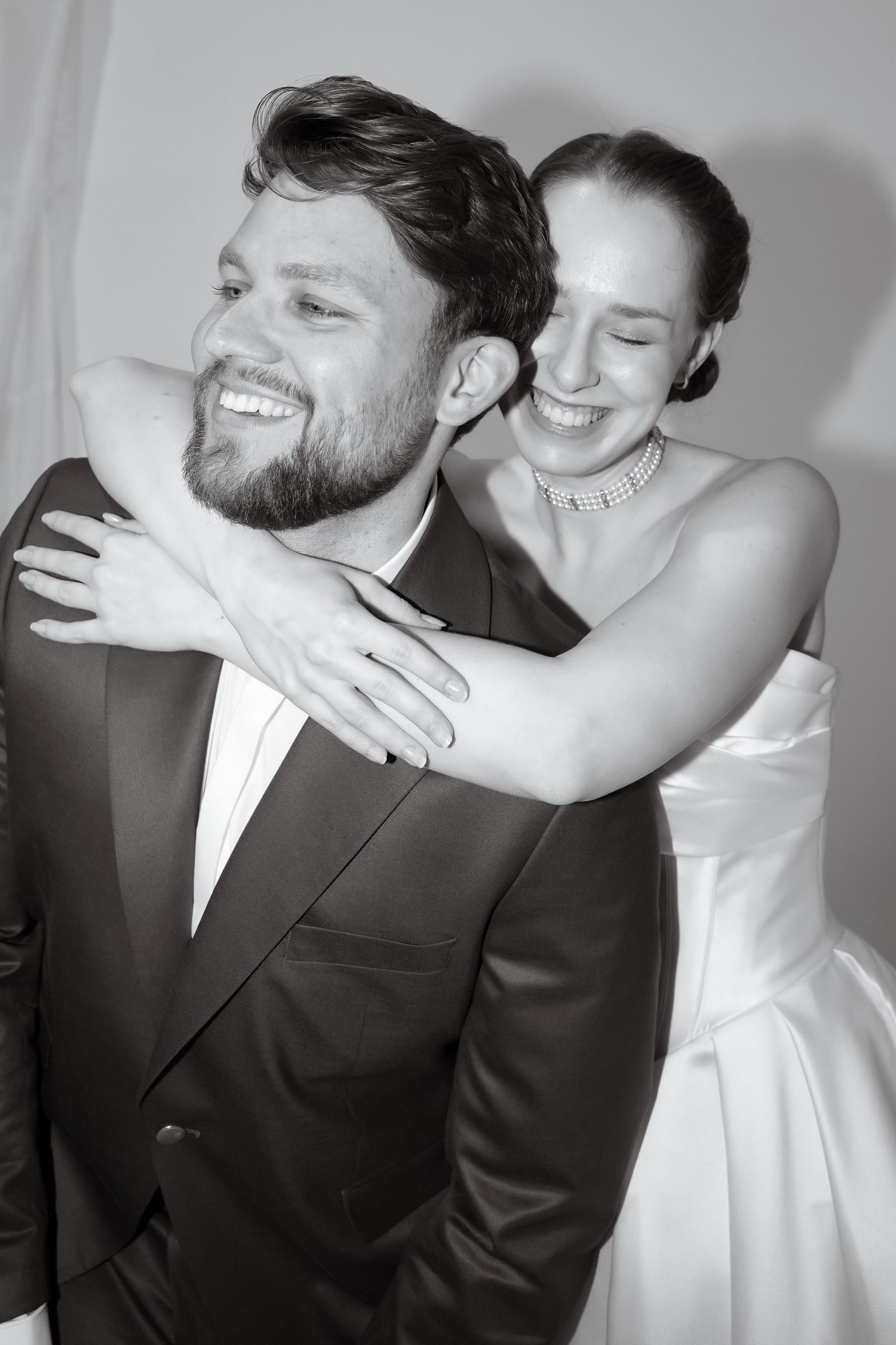 Black and white portrait of a smiling bearded groom in a suit as his bride in a strapless gown and pearl necklace hugs him from behind.