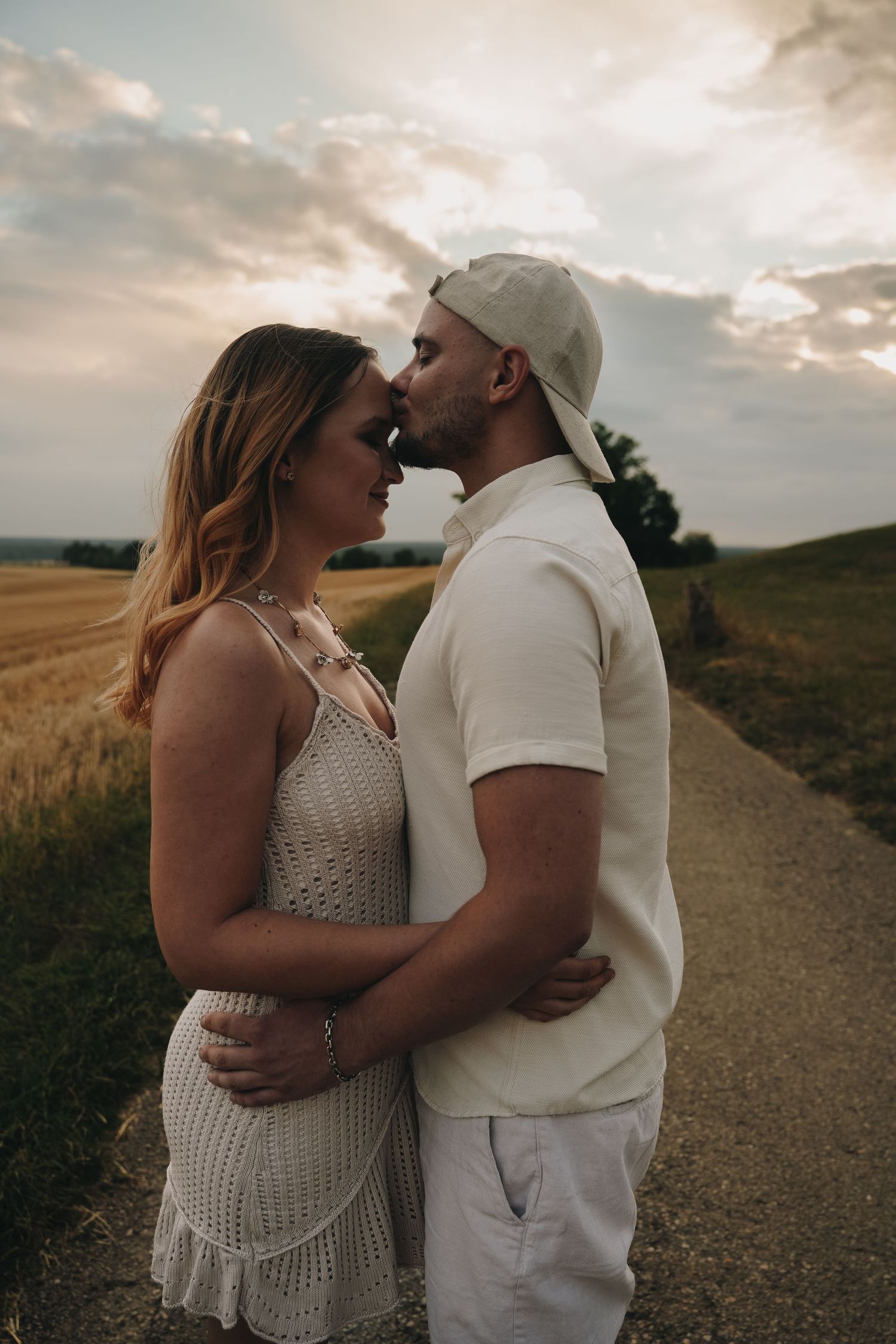 Couple sharing a romantic kiss on a countryside road at sunset, surrounded by golden wheat fields.