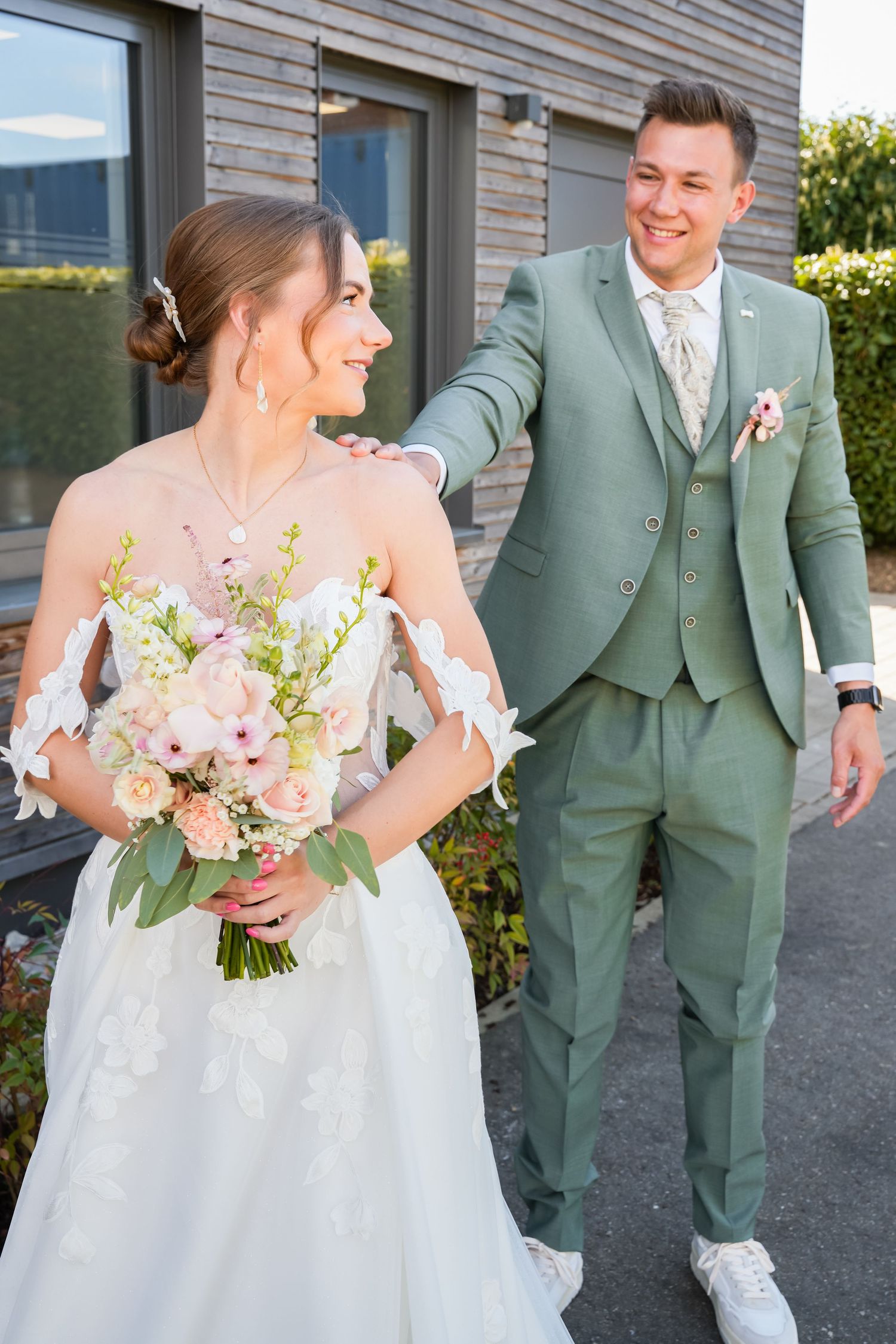 A smiling groom in a sage green suit places his hand on his bride's shoulder as she holds a pink bouquet.