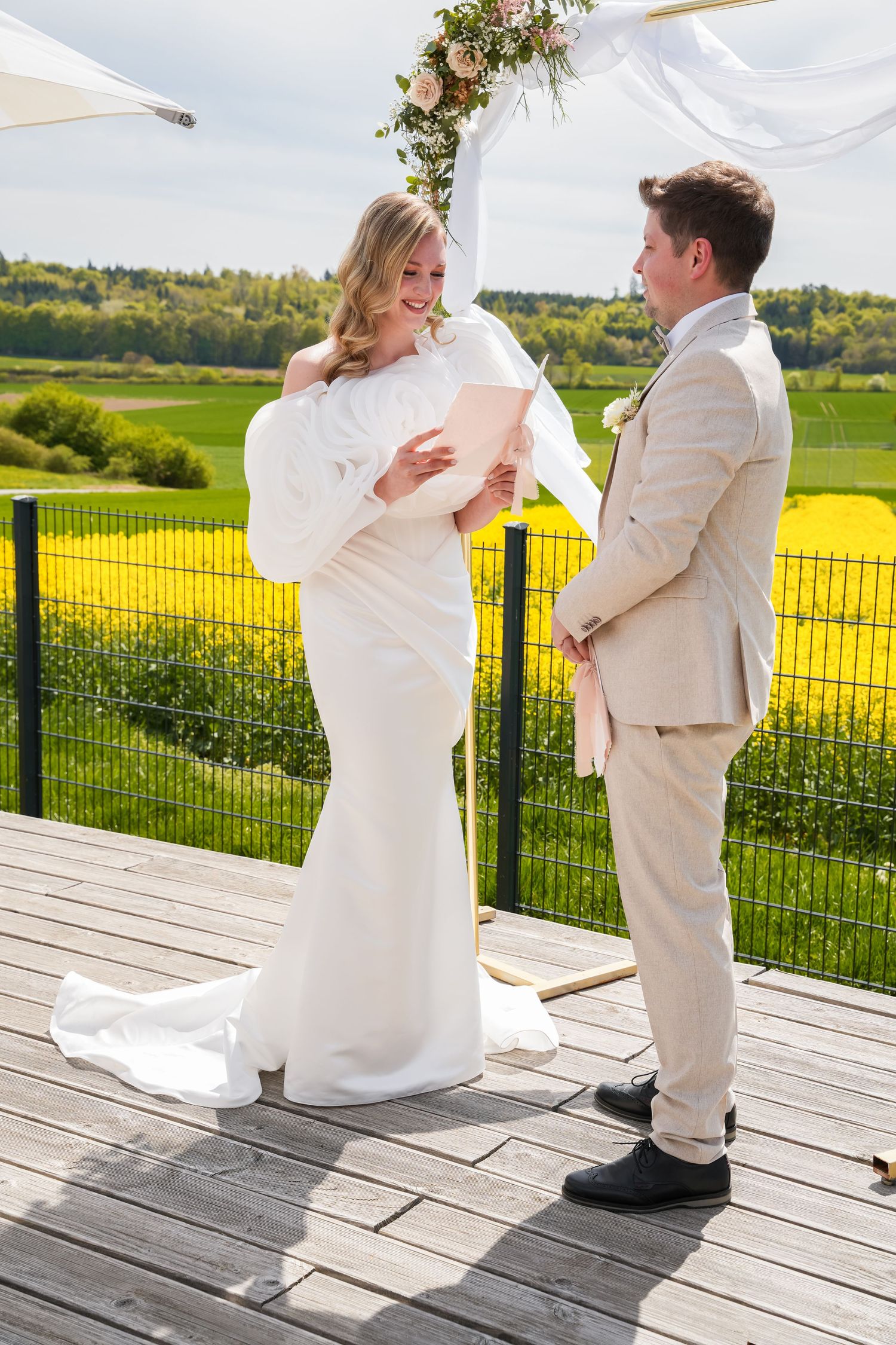 Bride in white gown reads vows to groom in beige suit on wooden deck with floral arch and yellow field backdrop.