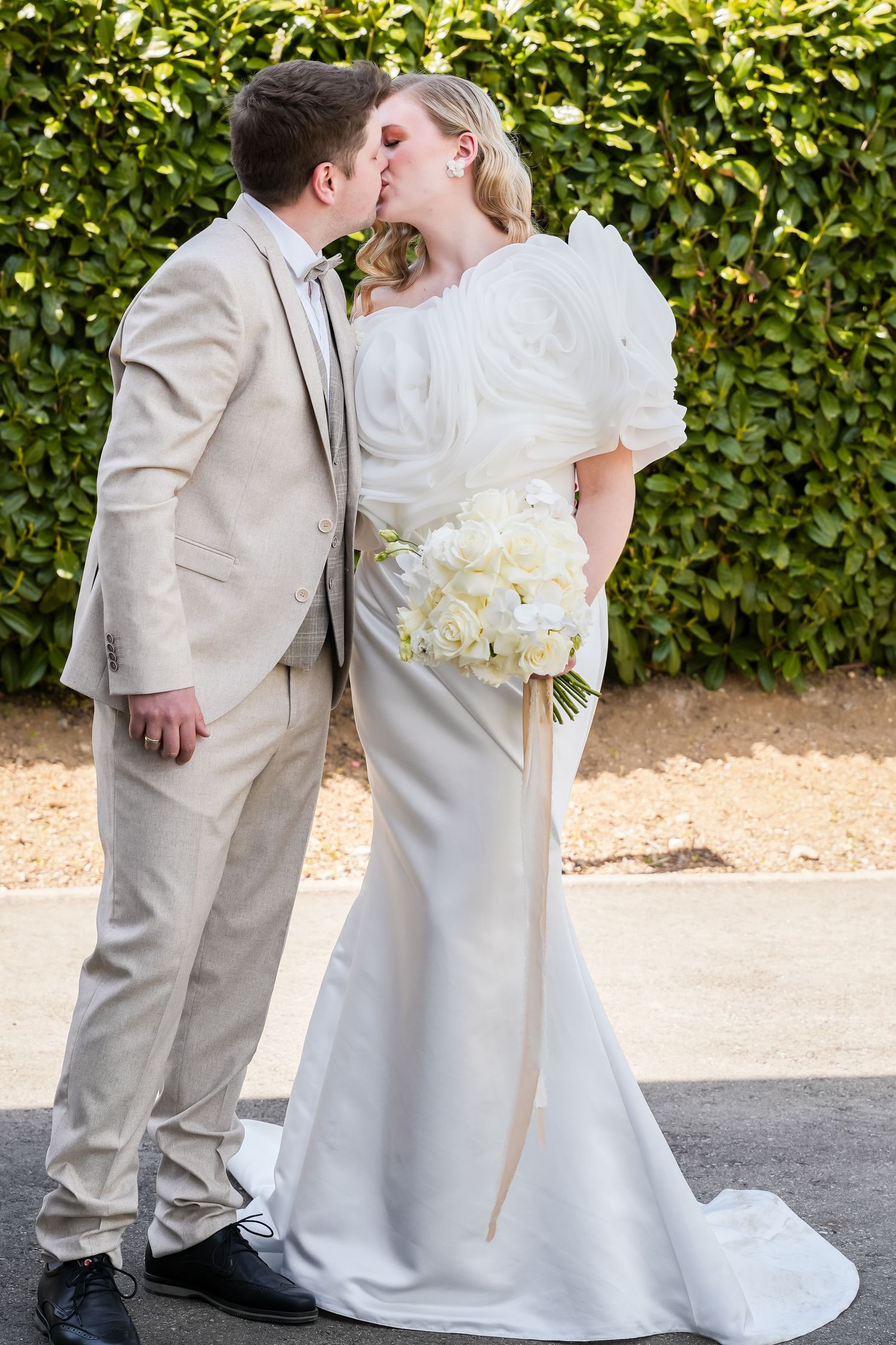 A bride and groom share a kiss outdoors, she holds white roses bouquet wearing satin gown, he wears beige suit.