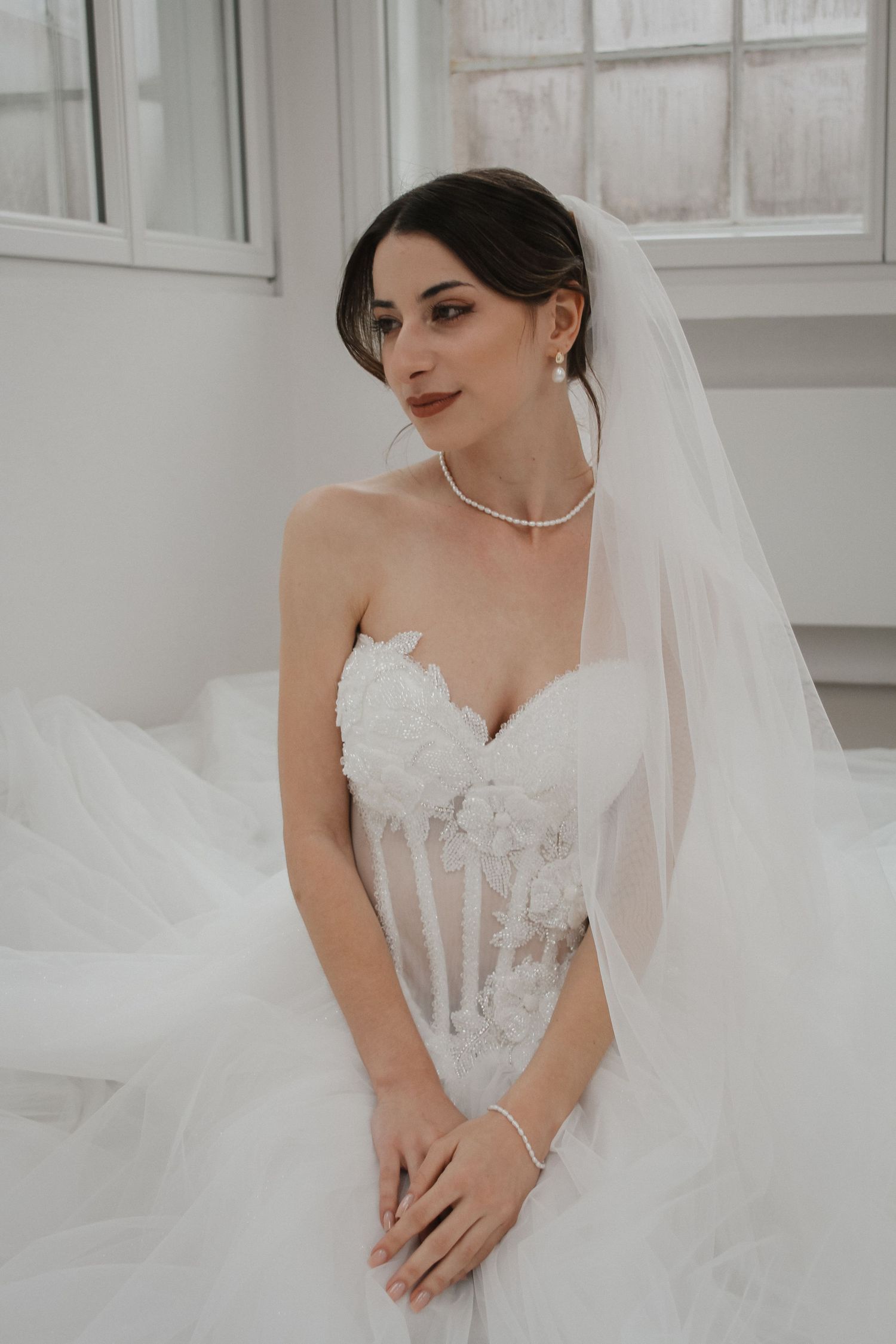 Elegant bride in strapless lace wedding gown with veil and pearl necklace posing gracefully indoors.