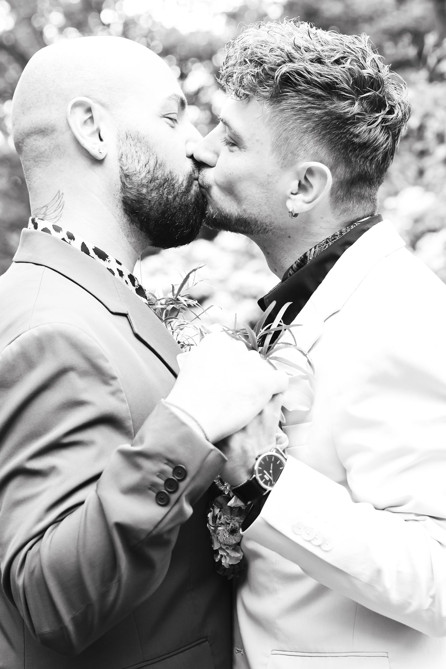 Black and white photo of two grooms sharing a kiss on their wedding day, both wearing suits with boutonnieres.