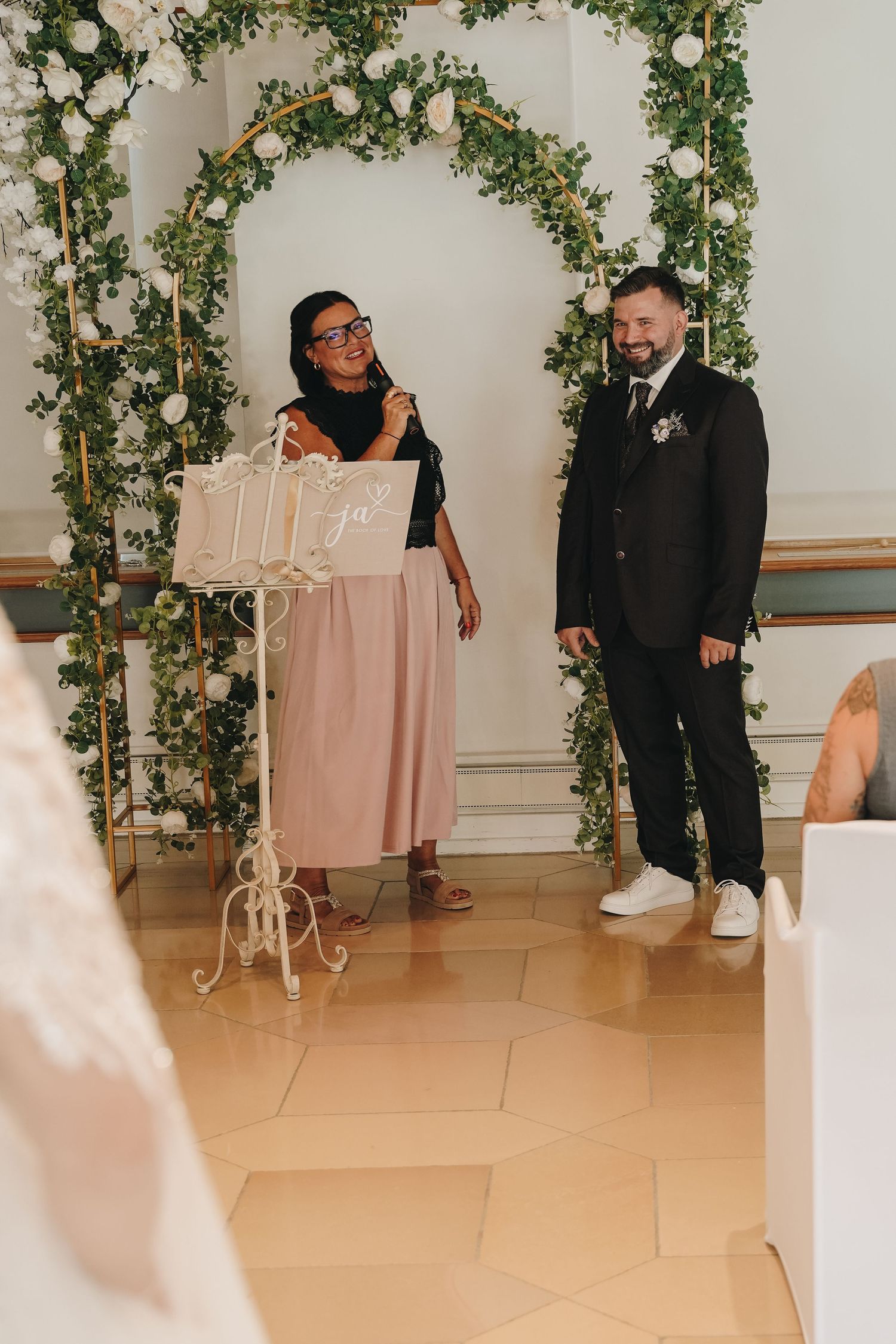 Smiling couple stands before a floral arch with greenery and white roses during an elegant wedding ceremony indoors.